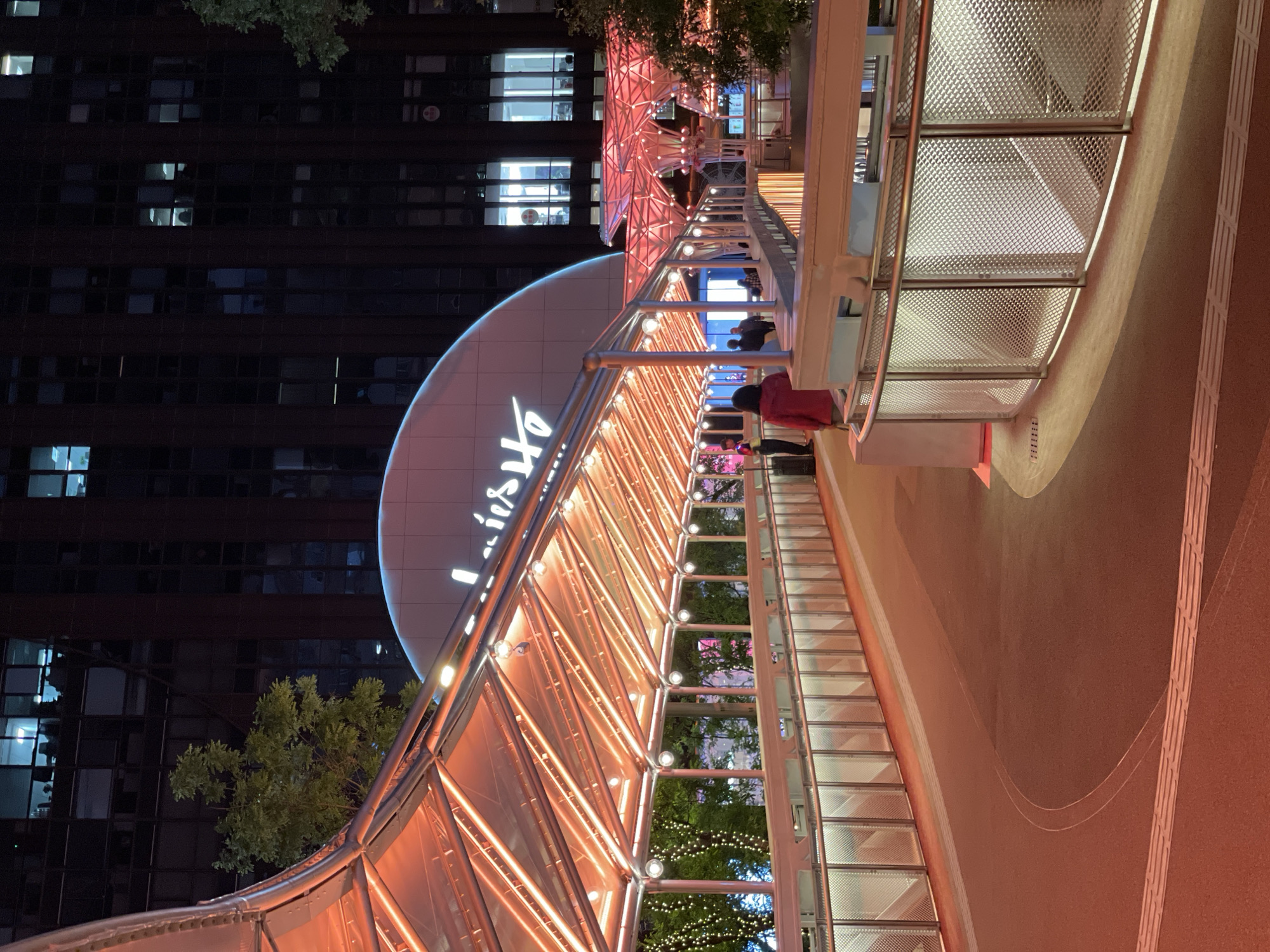 The image shows an illuminated, elevated walkway in Xicheng District, China, at night. The walkway has a steel frame structure with orange lighting, and metal mesh side panels. Pedestrians are visible on the walkway. In the background, there is a tall building with multiple illuminated windows and a large sign with the word "Lifestyle" in script. Some trees are visible along the walkway and in the background.