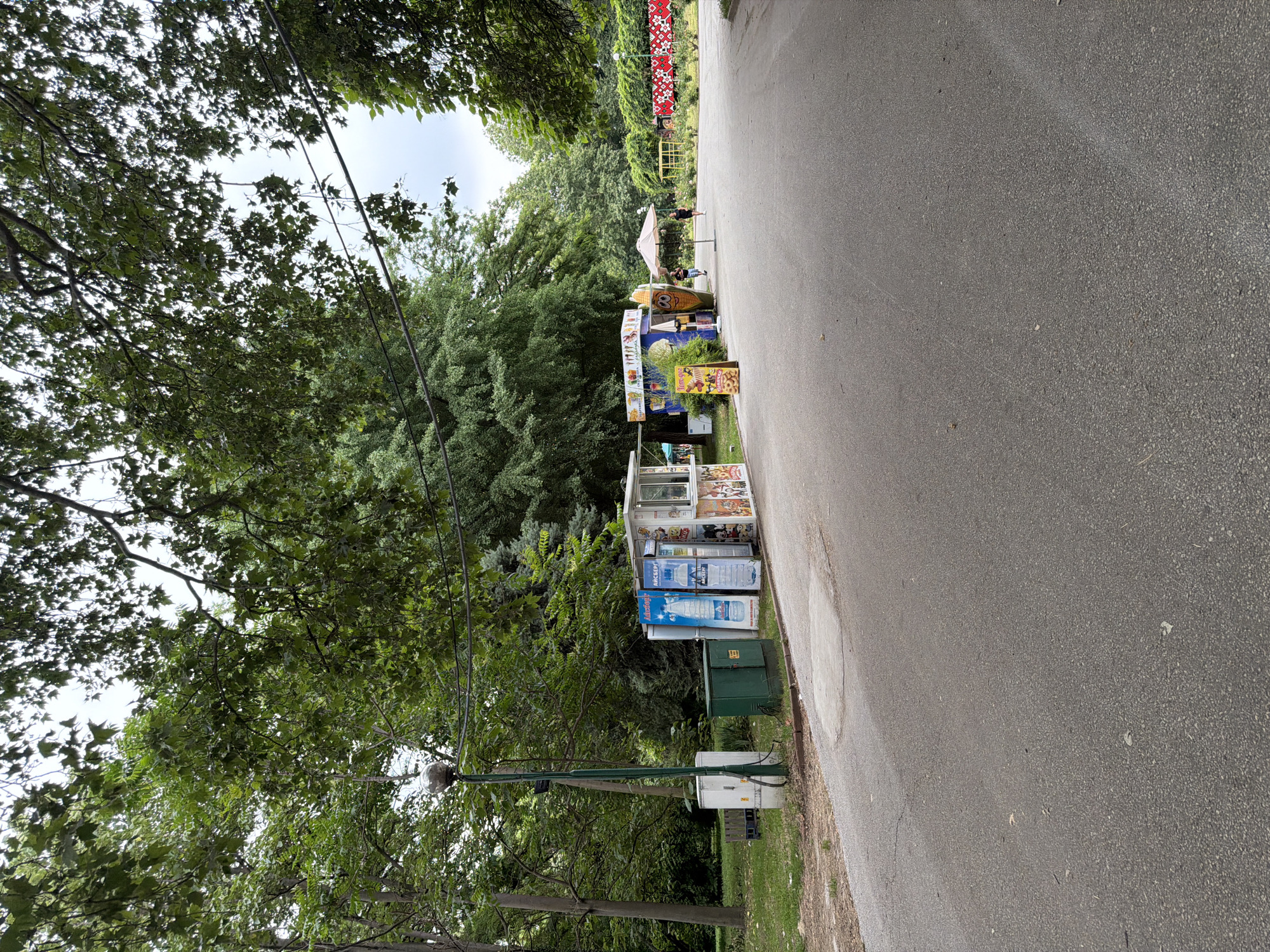 The image shows a paved pathway in a park with several small kiosks selling drinks and snacks. In the foreground, the pathway is mostly empty. To the left of the pathway, there are several kiosks, one with large bottles of water, another with an ice cream display. A lamppost stands in front of these kiosks. Beyond the kiosks, the pathway curves to the right and continues into the distance. In the mid-ground, there are more kiosks, one with a prominent corn on the cob decoration and an umbrella. Two people are walking on the pathway. In the background, there are many trees and lush greenery, creating a park-like setting. A decorative red and white patterned fence is visible on the right side of the pathway in the far background. The sky is overcast. The scene appears to be in a public park, likely an area within the city of Pazardzhik, Bulgaria, given the presence of these types of small food and drink vendors commonly found in Bulgarian parks and recreational areas.