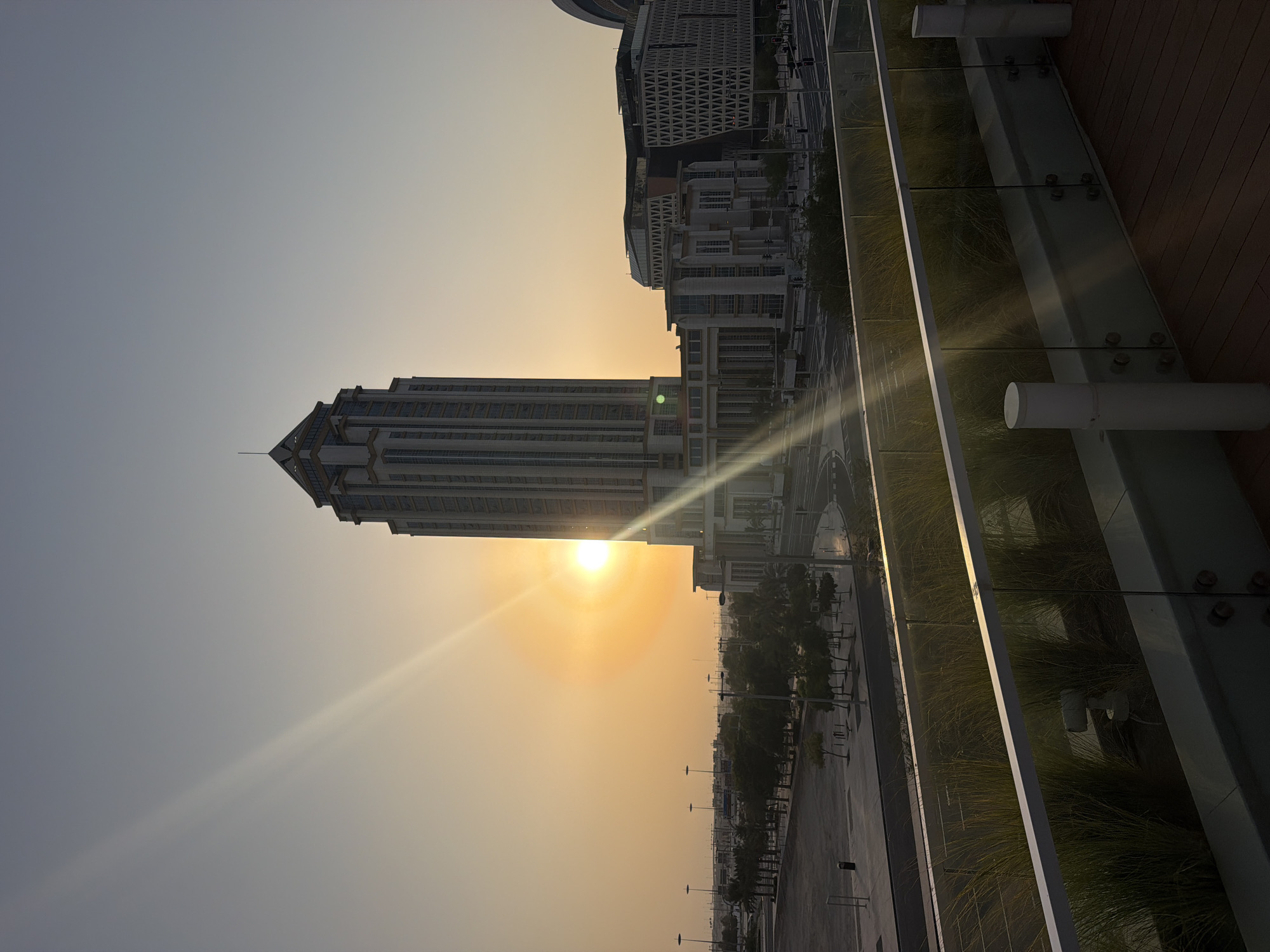 This image shows the Lusail cityscape in Qatar during sunset. A tall, modern skyscraper dominates the center of the frame, with a large, glowing sun positioned to its left, casting long rays of light across the scene. Several other contemporary buildings of varying heights and architectural styles are visible in the background, along with streetlights and rows of trees lining a road. In the foreground, there is a wooden deck with a glass railing, behind which is a planter filled with tall, green grass. The sky is a pale orange and grey, indicative of the twilight hour. There are no people visible in the image.