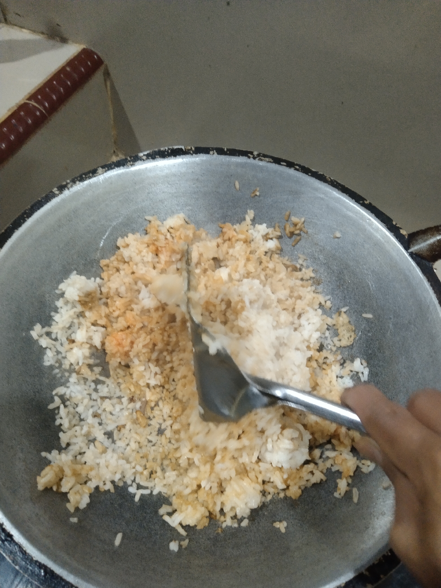 In the city of Suak Lanjut, Indonesia, a close-up shot captures a person's hand stirring fried rice in a large, worn wok. The rice is a mixture of white and lightly browned grains, suggesting it's in the process of being cooked or seasoned. A metal spatula is partially submerged in the rice, actively mixing the contents. The wok rests on what appears to be a stovetop, though only a portion of the rim is visible. The background is blurred and nondescript, with muted tones of beige and grey, possibly indicating a kitchen wall or counter. The overall scene conveys a sense of domestic cooking.