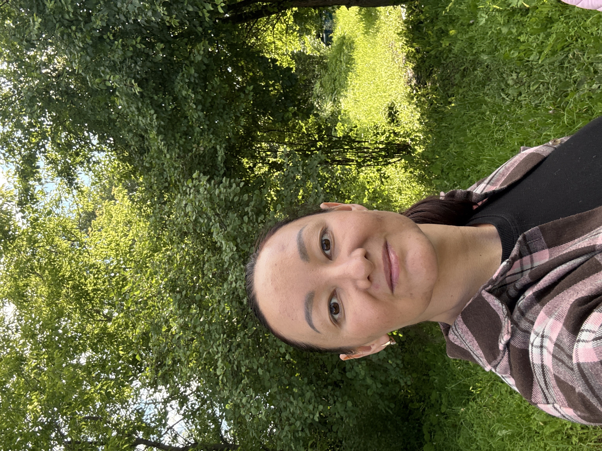 A woman with dark hair and brown eyes takes a selfie outdoors in a natural setting. She is wearing a pink and brown plaid shirt over a black top. The background consists of lush green trees and grass, with sunlight filtering through the leaves, creating a dappled effect. The location is the village of Bolshoy Turysh, Russia.