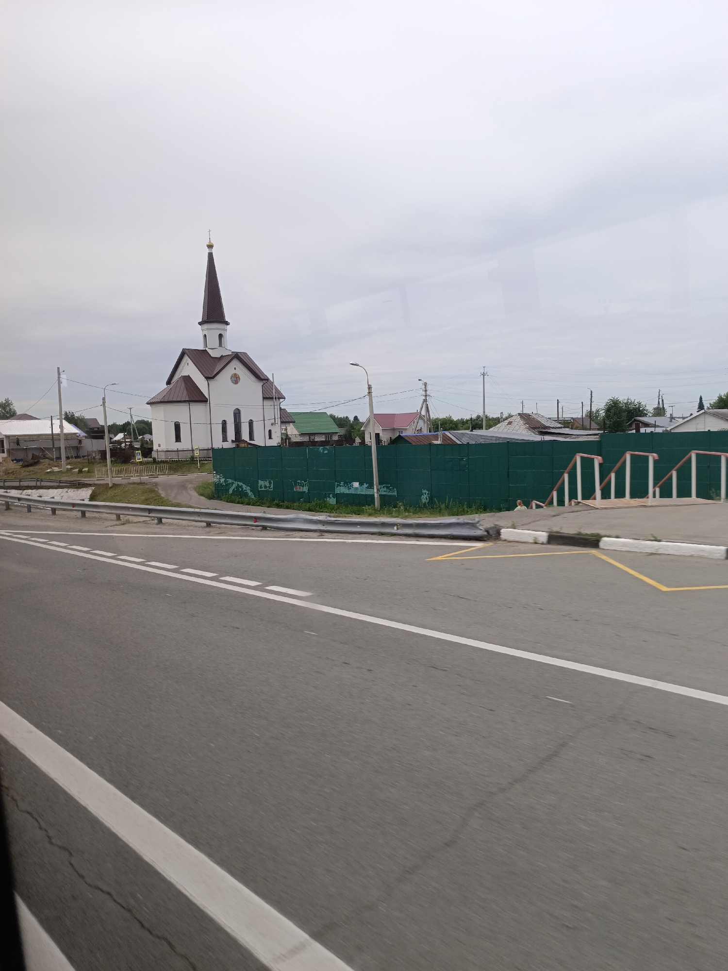 In Zudilovo, Russia, a white church with a brown, pointed steeple topped with a cross stands to the left of a bright green metal fence. The church has a brown roof and two rows of windows. The fence runs horizontally across the middle of the image, separating the church and surrounding buildings from a grey asphalt road in the foreground. White dashed lines mark the lanes on the road, and a white curb with yellow markings is visible. Beyond the fence, other buildings with varied roof colors and styles are scattered. The sky is overcast and grey.