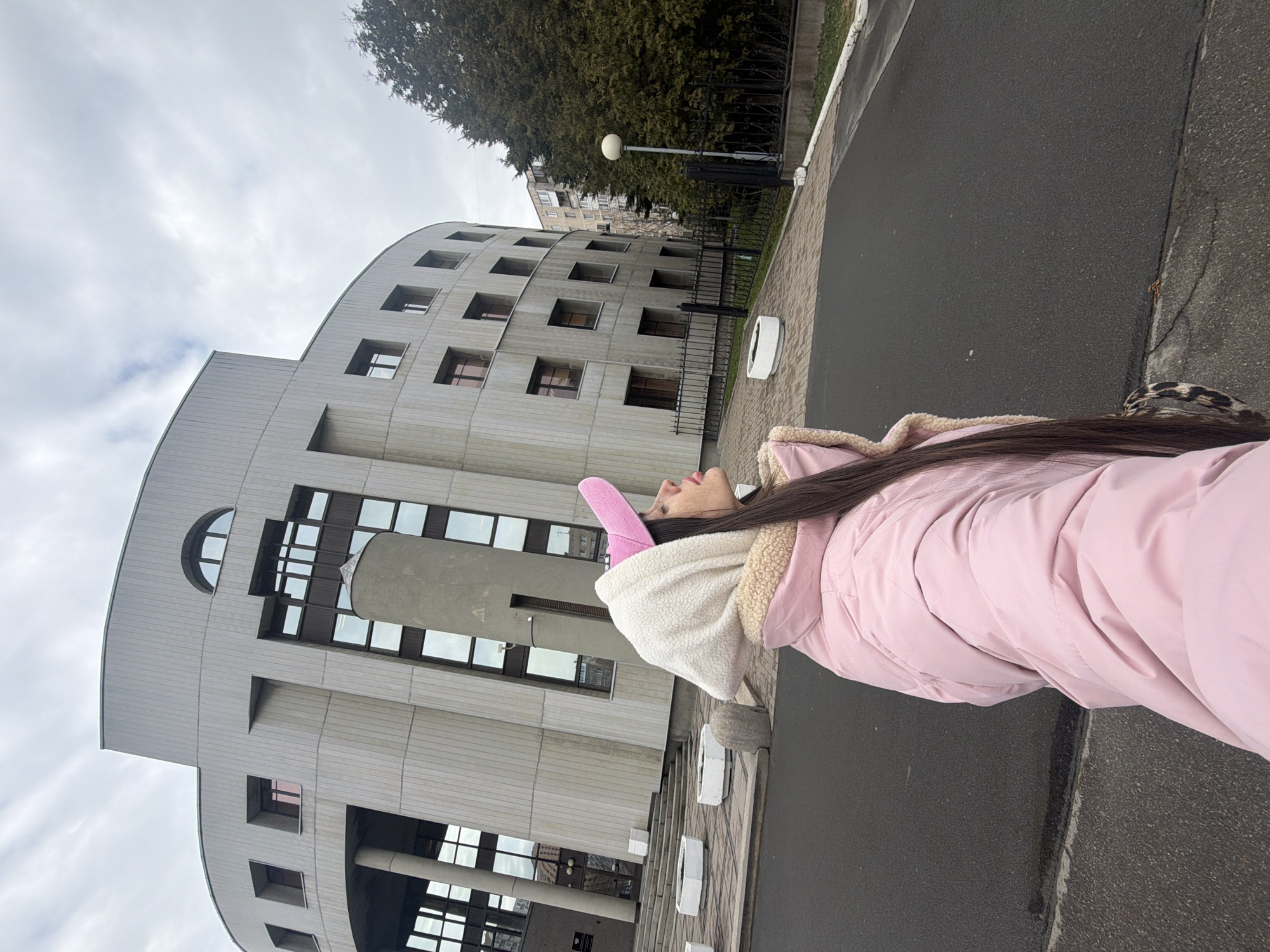 A vertical, eye-level shot depicts a woman in a pink jacket and pink hat standing on a paved surface with a large, grey building in the background.
The woman is on the right side of the frame, facing left. She is wearing a light pink jacket and a white hooded sweater with a beige fleece lining. A pink hat is on her head. Her hair is long and dark.
The building behind her is a large, grey, modern-style structure with multiple square windows and curved sections. A lamppost and trees can be seen behind the building, along with other buildings in the distance. The sky is overcast.
Based on the architectural style of the building, this appears to be the Central Naval Library located in Saint Petersburg, Russia.