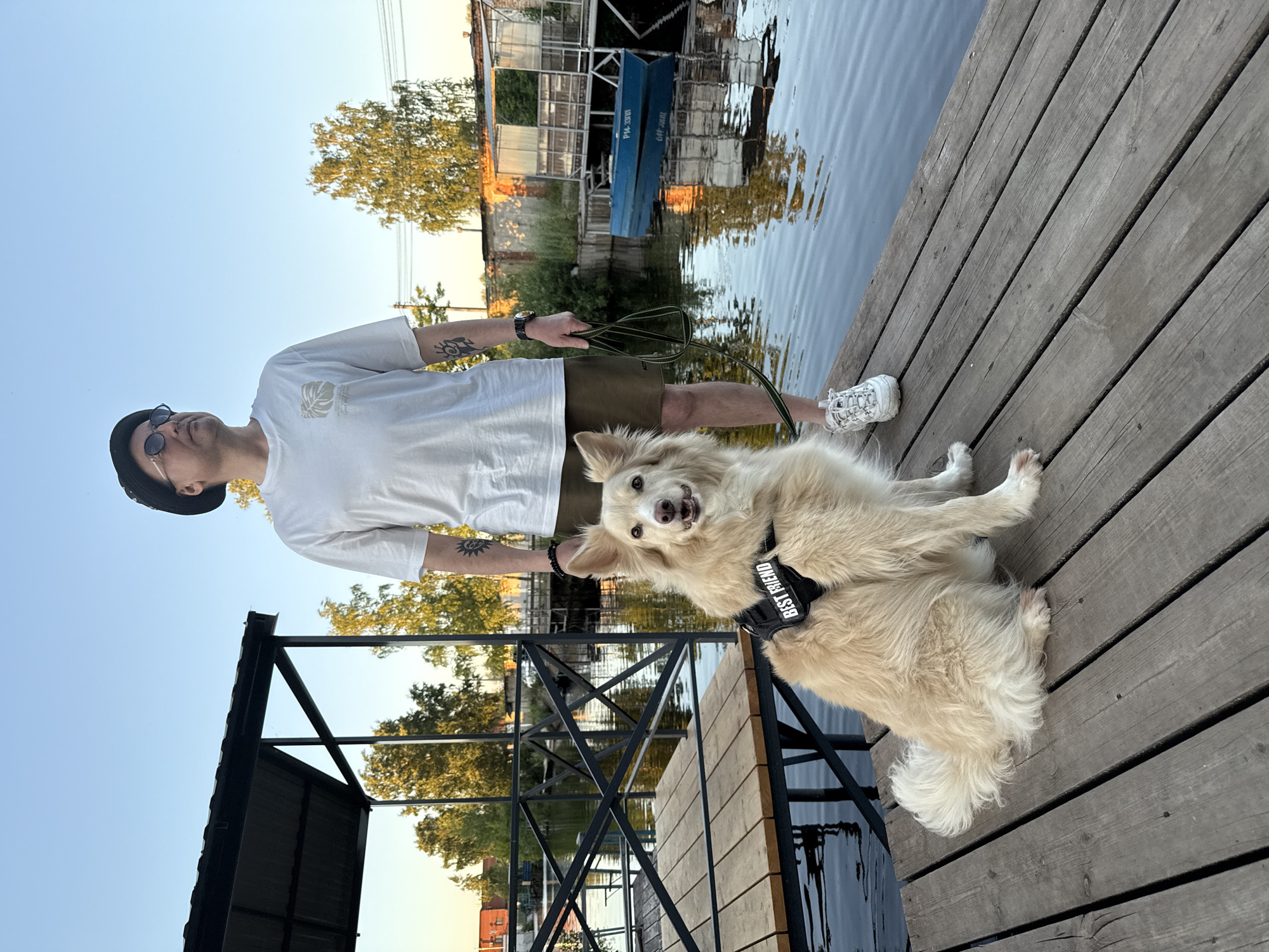 A man in a white t-shirt, olive shorts, sunglasses, and a black hat stands on a wooden pier next to a light-colored dog. The dog is wearing a black harness with "BEST FRIEND" written on it. The man holds a leash in his right hand. In the background, there is a body of water, trees, and buildings. A blue boat is visible on the water. The scene appears to be in Chaikovsky, Russia, likely near the Kama River. The sun is low in the sky, suggesting it is either sunrise or sunset.