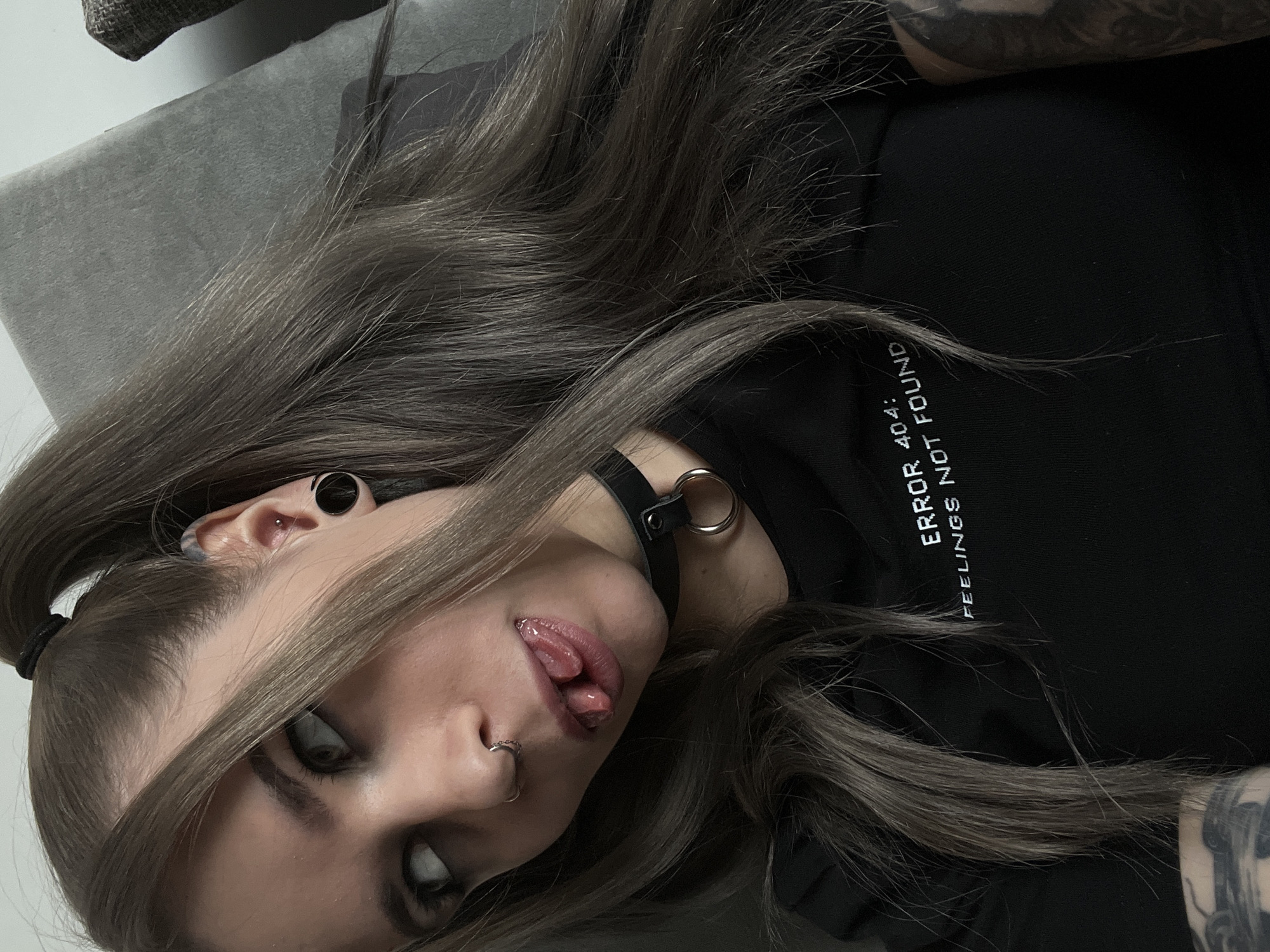 A young woman with long, gray-brown hair is lying on a gray surface, possibly a sofa or bed. Her head is tilted, and she is sticking out her tongue. She has several piercings, including a nose ring and gauged ears. She is wearing a black choker with a metal ring and a black t-shirt with the text "ERROR 404: FEELINGS NOT FOUND". Tattoos are visible on her arm. The background appears to be a simple, plain wall. There is no visible information in the image to determine if the location is Khimki, Russia.