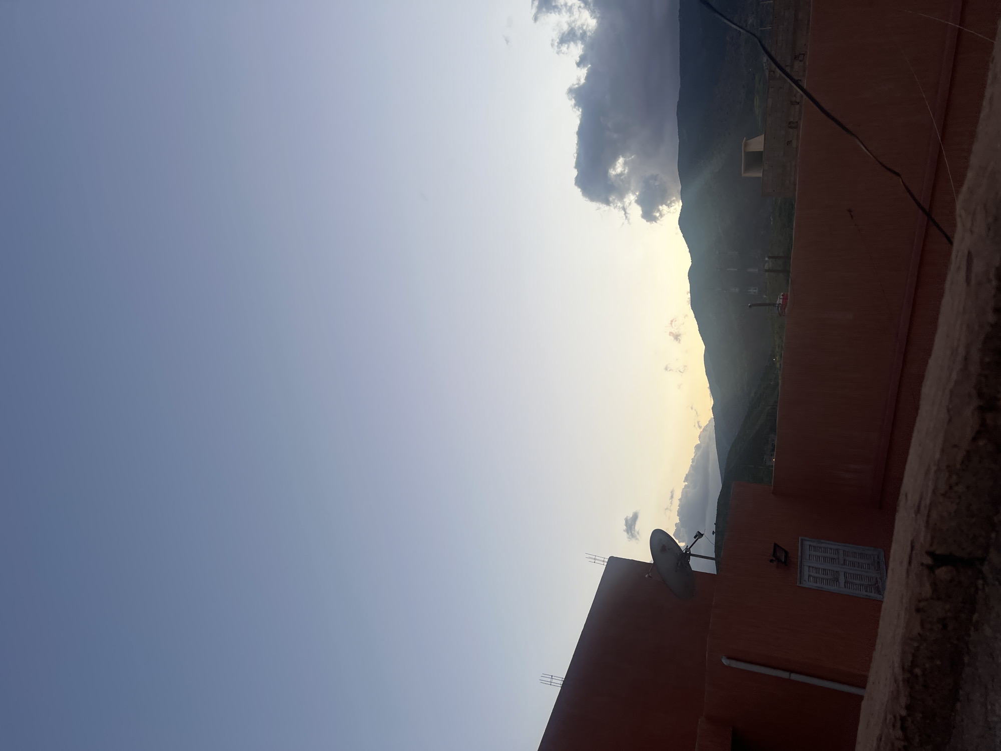 The image depicts a view from what appears to be a rooftop in Ouaouizert, Morocco. A building with a satellite dish and a window is visible in the foreground. In the background, there is a mountain range partially obscured by clouds. The sky transitions from light blue to a yellowish hue near the mountains, suggesting either sunrise or sunset.
