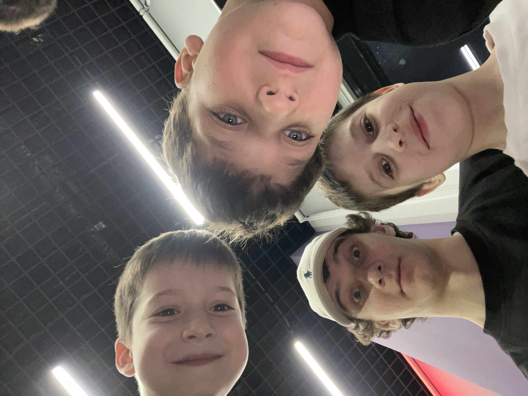 A low-angle, wide-lens photograph depicts four male individuals looking downwards into the camera within an indoor commercial or recreational environment.
In the upper left quadrant, a male child, estimated 8-12 years old, with short brown hair and dark eyes, shows a slight smile. To his right and slightly lower, another male child, approximately 10-14 years old, with short brown hair and blue eyes, maintains a neutral, direct gaze. In the lower left, a male young adult, likely late teens to early twenties, wears a white baseball cap with a dark blue polo player logo, and a black t-shirt. He has curly brown hair and wide, open eyes, conveying a surprised or playful expression. To his right, partially obscured by the young adult, a third male child, estimated 8-12 years old, with short brown hair and dark eyes, smiles while wearing a white t-shirt.
The background features a dark, gridded ceiling, indicative of a suspended tile system. Multiple linear fluorescent or LED light fixtures emit bright white light, with one diagonal fixture prominent in the upper left. A segment of a purple-pink wall is visible in the lower-left background, and a white panel with a metallic rail is present on the right. The scene's specific location within Krasnoyarsk, Russia, cannot be definitively identified from the visual information provided but is consistent with a modern indoor public space such as a shopping mall or entertainment complex.
