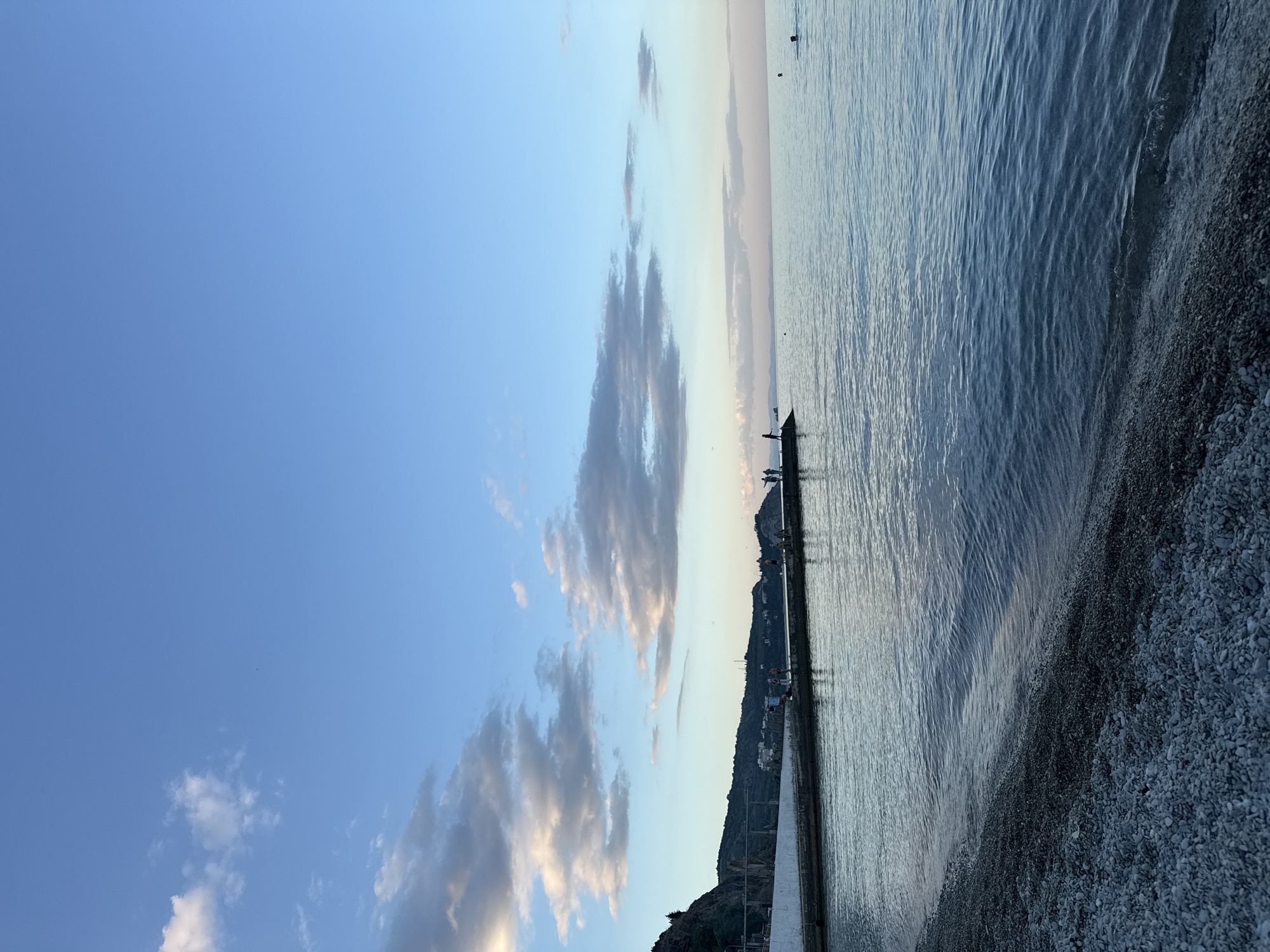 The image depicts a serene seascape in Partenit, Ukraine, likely at dusk. A pebble beach occupies the foreground, with the gentle waves of the sea lapping at its edge. In the mid-ground, a concrete pier extends into the water, populated by a small group of figures, some appearing to be fishing or observing the view. Further out in the water, a lone swimmer can be seen. The background features rolling hills and a coastal town, with a vast, clear sky above, partially filled with scattered clouds. The overall impression is one of tranquility and natural beauty.