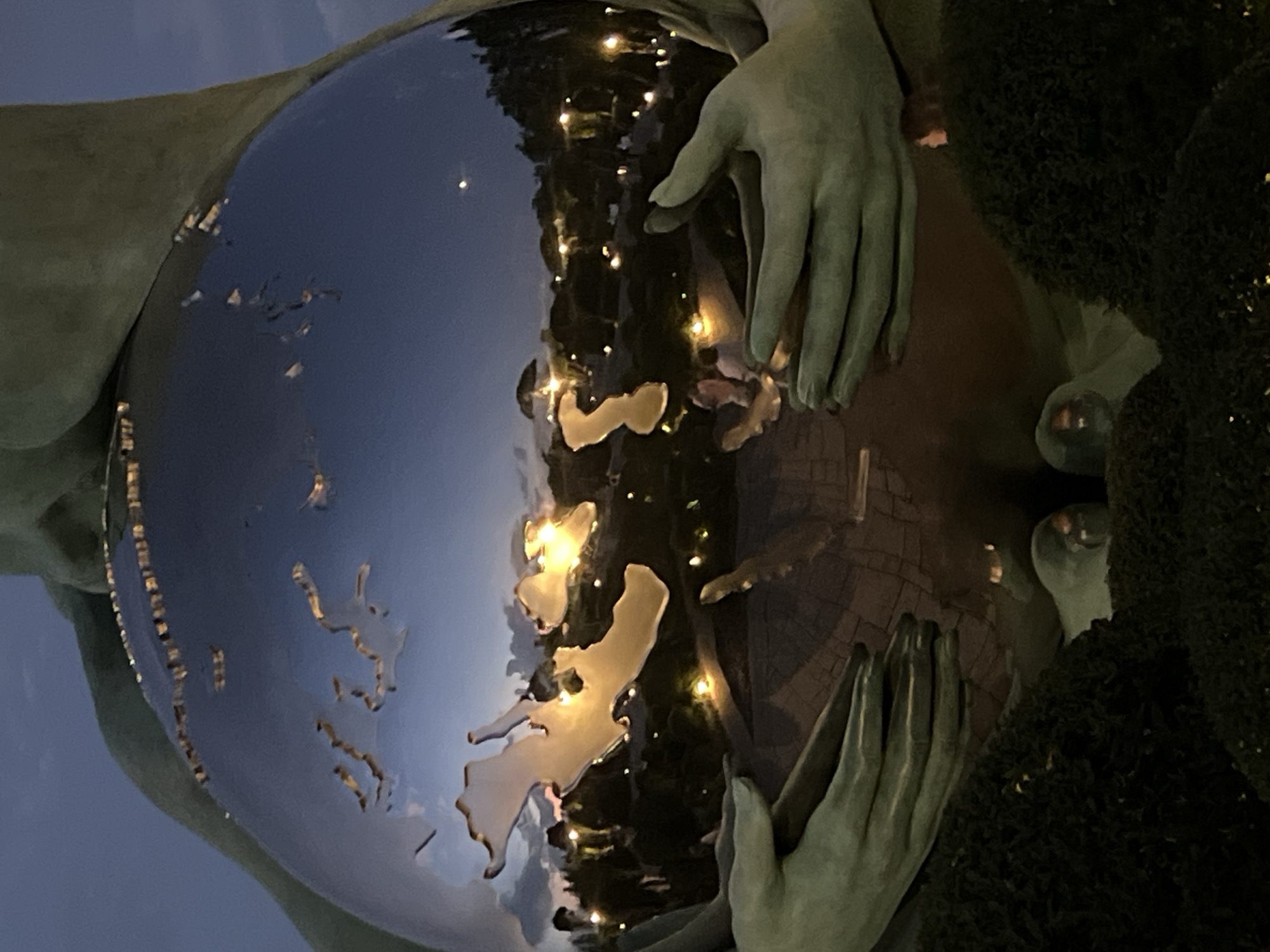 This media file captures a close-up of a sculpture, likely outdoors at dusk, given the twilight sky visible in the reflection. The sculpture features large, stylized hands gently cradling a spherical object. This sphere appears to be a representation of the Earth, with the continents etched into its surface. The surface of the sphere is highly reflective, mirroring the surrounding environment.
Visible in the reflection are numerous small, bright lights, suggesting the presence of streetlights or decorative lighting. There are also dark, shadowy areas reflecting, which could be trees or structures. The hands of the sculpture are rendered in a muted, greenish-gray tone, giving them an aged or weathered appearance. One hand is positioned on the left side of the sphere, while another is on the right, both appearing to hold the Earth protectively.
The overall mood conveyed is one of care, guardianship, and possibly a sense of wonder or contemplation towards the planet. The orientation of the image is rotated, presenting a dynamic and unusual perspective. The location is identified as Krasnodar, Russia, but specific landmarks are not identifiable within the current frame or its crops. No visible text is present in the image.