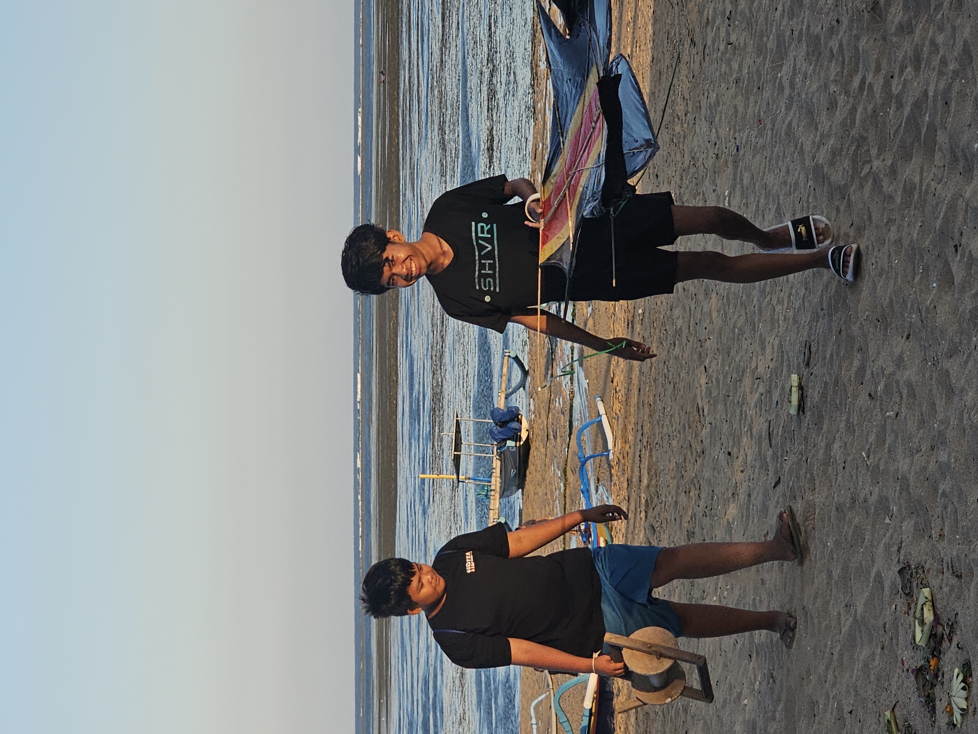 Two young males are walking on a beach in Denpasar, Indonesia, with the ocean and sky in the background. The male on the left is wearing a black t-shirt and blue shorts. He is carrying a large wooden spool with string on it. The male on the right is wearing a black t-shirt and black shorts. He is holding a large, colorful kite. Several small boats are visible on the beach and in the shallow water. The sand is light brown and appears wet in some areas. The sky is a clear, pale blue.