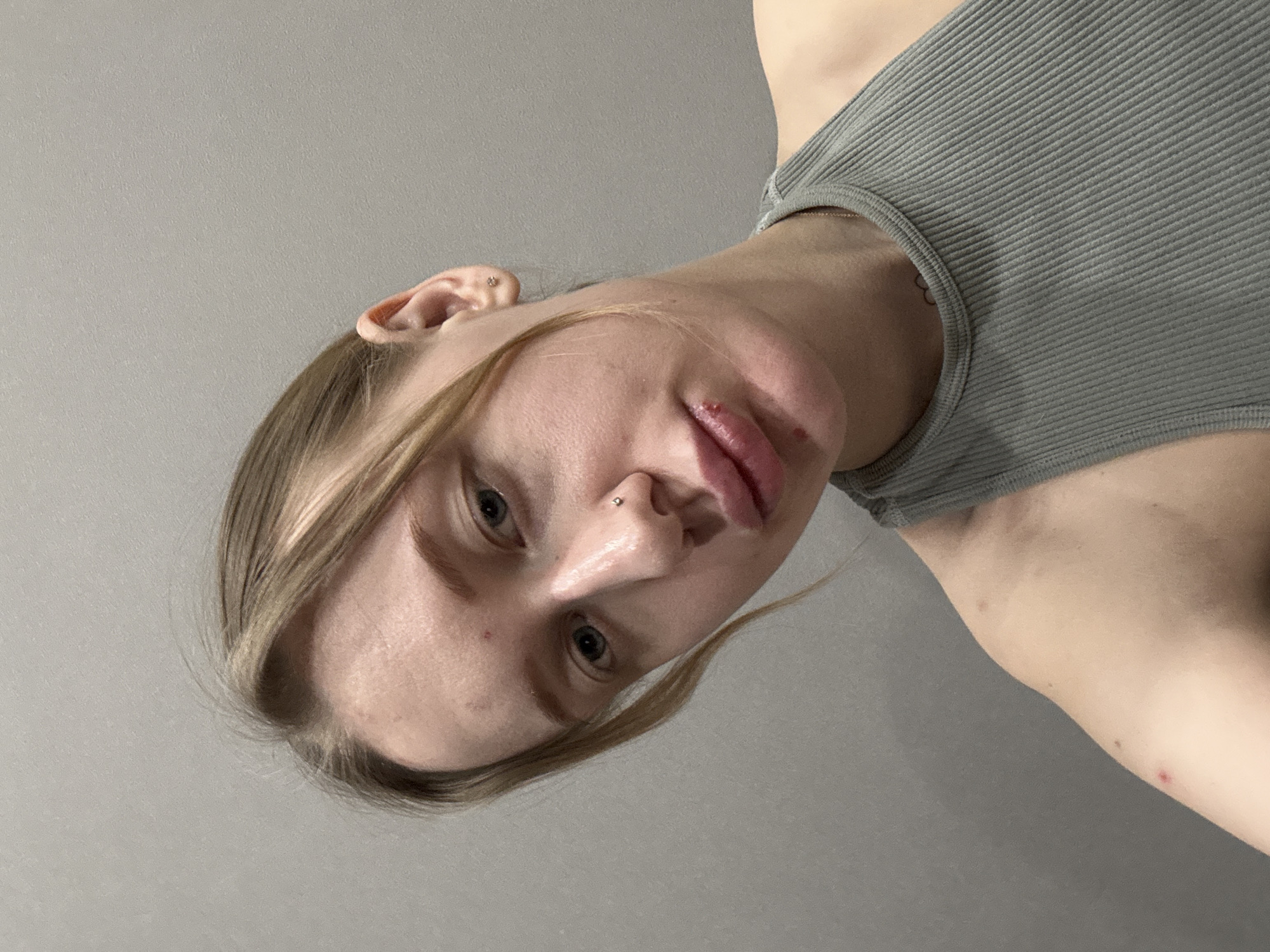 A close-up portrait depicts a fair-skinned individual with blonde hair, seemingly indoors against a grey background. The subject's face is slightly tilted, revealing light blue eyes, a nose piercing, and a mark on the lip. The person is wearing a grey ribbed tank top. The environment is unremarkable, with only the wall visible. There's no indication of this being in Rostov-on-Don.