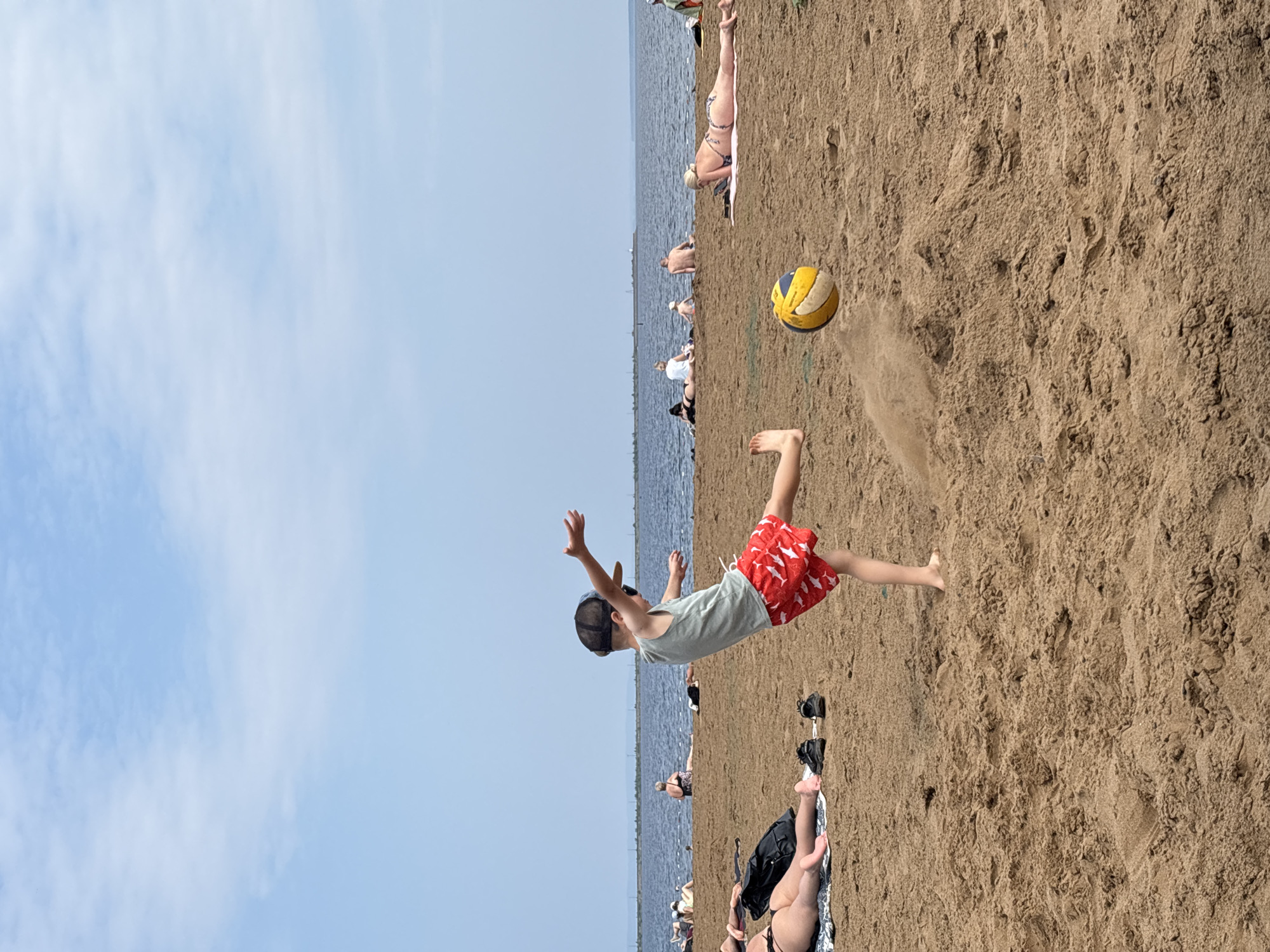 The image depicts a sunny day at a sandy beach, likely on the Kama River embankment in Chaikovsky, Russia, given the context. A young boy, wearing a light grey tank top, red shorts with a white shark pattern, and a dark baseball cap, is captured in mid-action, kicking a volleyball. The ball, colored yellow and blue, is in the air, with a puff of sand kicked up by the boy's foot. Several other people are scattered across the beach, some lying down or sitting, enjoying the weather. In the background, the calm, wide expanse of the Kama River stretches out, with a thin strip of land visible on the opposite bank. The sky is a pale blue with scattered white clouds, indicating a clear day. The overall atmosphere is one of leisure and recreation.