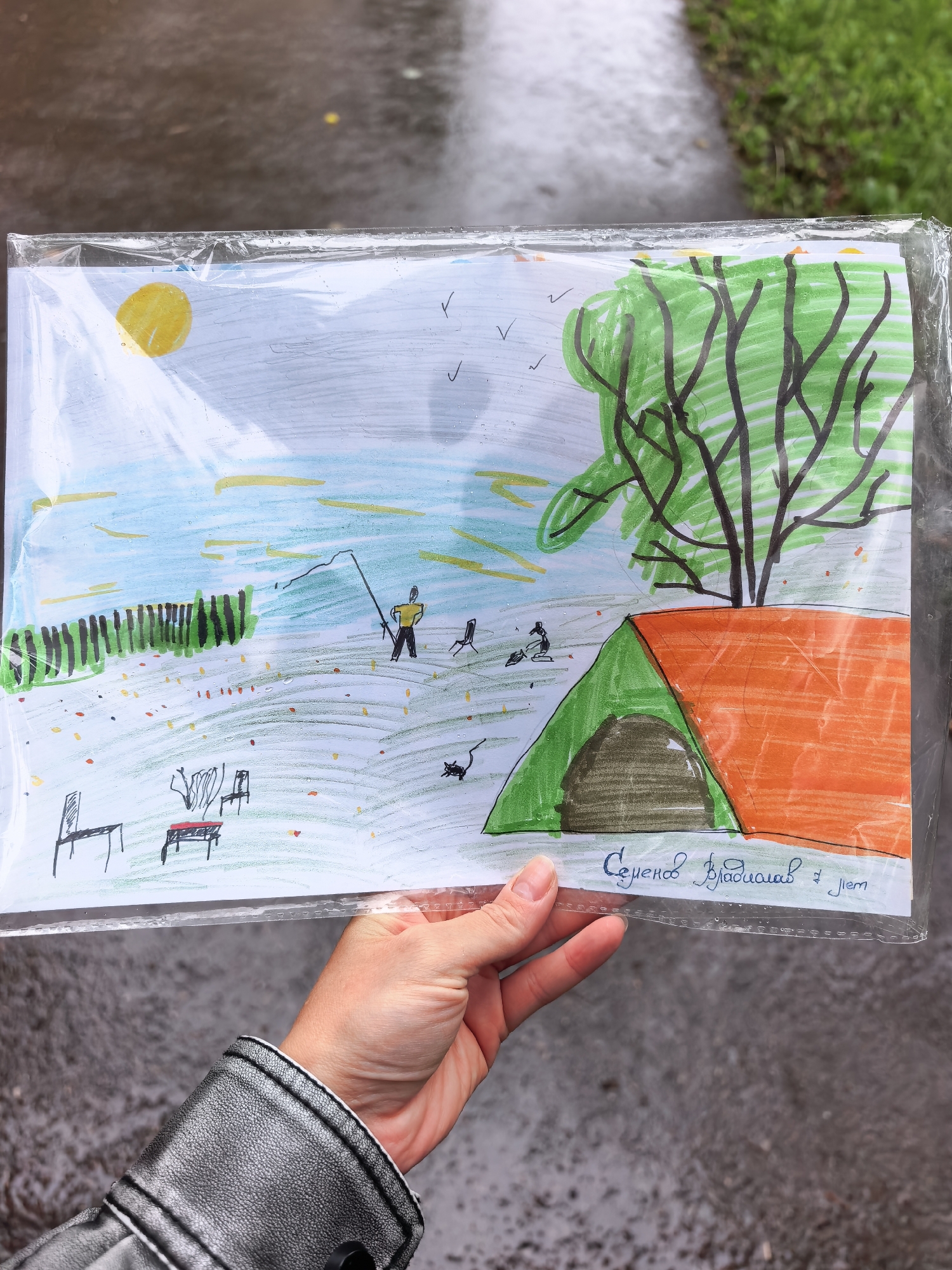 The image displays a child's drawing, enclosed in a clear plastic sleeve, held by a person's hand. The drawing depicts a vibrant outdoor camping and fishing scene under a bright yellow sun in a light blue sky, with a few birds flying.
The main subjects in the drawing include a person in a yellow shirt fishing with a rod near a body of blue water. Along the water's edge, there's a dark green fence-like structure. In the foreground, a grassy area dotted with colorful specks suggests either flowers or fallen leaves. To the right, a large tree with a green canopy and black branches stands next to an orange and green camping tent. Further to the left, a small campfire is depicted with two chairs and a table, and a small black cat is also visible. Another small figure appears to be near the campfire.
The activity taking place in the drawing is an idyllic outdoor recreational scene, combining fishing and camping. The time of day is clearly daytime, indicated by the sun. The weather in the drawing appears pleasant. However, the real-world setting of the photo suggests different conditions: the plastic sleeve has water droplets, and the blurry background shows wet, dark pavement and green grass, indicating it is raining or has recently rained outdoors in Sterlitamak, Russia. The drawing is childlike and uses vivid colors.
Visible text on the drawing, handwritten in blue script at the bottom right, reads: "Семенов Владислав 7 лет," which translates to "Semenov Vladislav 7 years old." The hand holding the drawing wears a grey, textured leather jacket with white stitching.