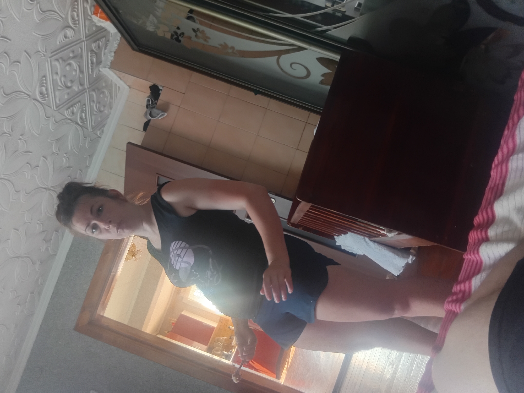 A woman in a black tank top and dark shorts stands in a room with patterned wallpaper. She is looking towards the camera with a neutral expression. Her hair is pulled back in a bun. A wooden table is to her right, and a large mirror is in front of her, reflecting a doorway and part of the room. The room appears to be indoors, possibly a home, with natural light streaming in from the doorway. The time of day is not explicitly indicated, but the lighting suggests it could be daytime.
