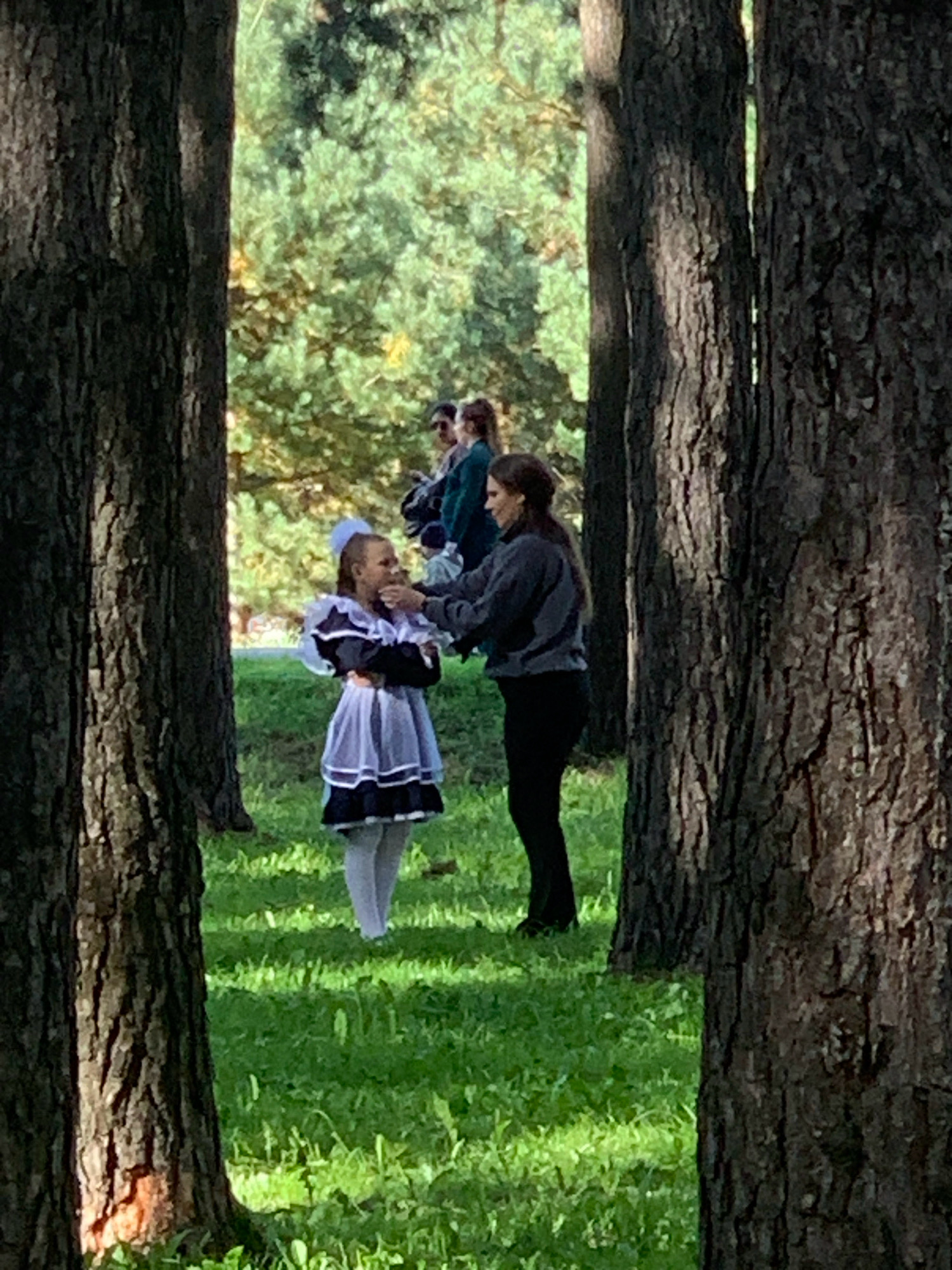 The image depicts a wooded area with two women in the foreground, framed by large trees. A young girl, dressed in a traditional school uniform with a white collar and blue skirt, stands on the left, facing a woman on the right. The woman on the right is wearing a dark gray sweatshirt and black pants. She is facing the girl and appears to be interacting with her, possibly adjusting her clothing or hair. In the background, several other individuals are visible, partially obscured by trees and foliage, suggesting a gathering or event. The setting is an outdoor park with lush green grass and trees that indicate a temperate climate. The overall impression is that of a staged or informal event taking place in a natural, outdoor urban environment. This scene is set in a park in the city of Prokopyevsk, Russia.