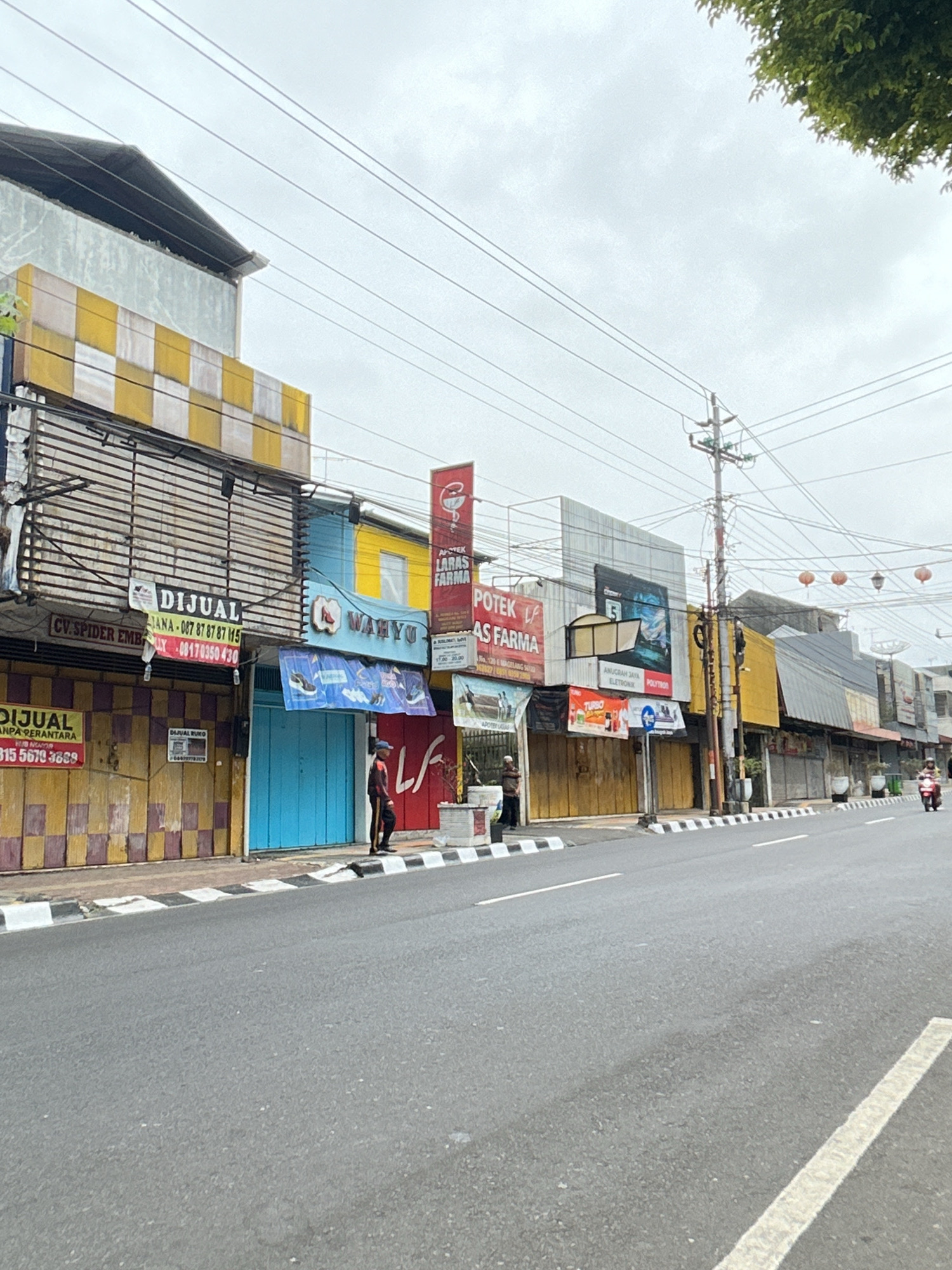 The image depicts a street scene in Magelang, Indonesia, characterized by a row of shopfronts with rolling shutters that are mostly closed. The architecture features multiple levels and a mix of colors, with prominent yellow and checkerboard patterns on some of the upper levels. Several signs are visible, including those advertising businesses like "DIJUAL" (for sale), "CV. SPIDER EMB," "WAHYU," "POTIK LARAS FARMA," and "AS FARMA." There are also signs for electronics businesses, with one displaying an image of a laptop.
In the foreground, a paved road with white lane markings runs diagonally across the frame. The sidewalk area in front of the shops is marked with black and white striped bollards and a curb. Two individuals are present on the sidewalk; one, wearing a dark uniform with an orange stripe, stands near a blue doorway. Another person, dressed in dark clothing, is further down the sidewalk, near a utility pole. A motorcycle is visible in the distance on the right side of the road, with a rider on it. The sky is overcast, contributing to a muted lighting condition. Overhead, power lines and a utility pole are prominent features of the environment. Red lanterns are hung above some of the buildings in the background. Lush green foliage from a tree is visible in the upper right corner of the image.