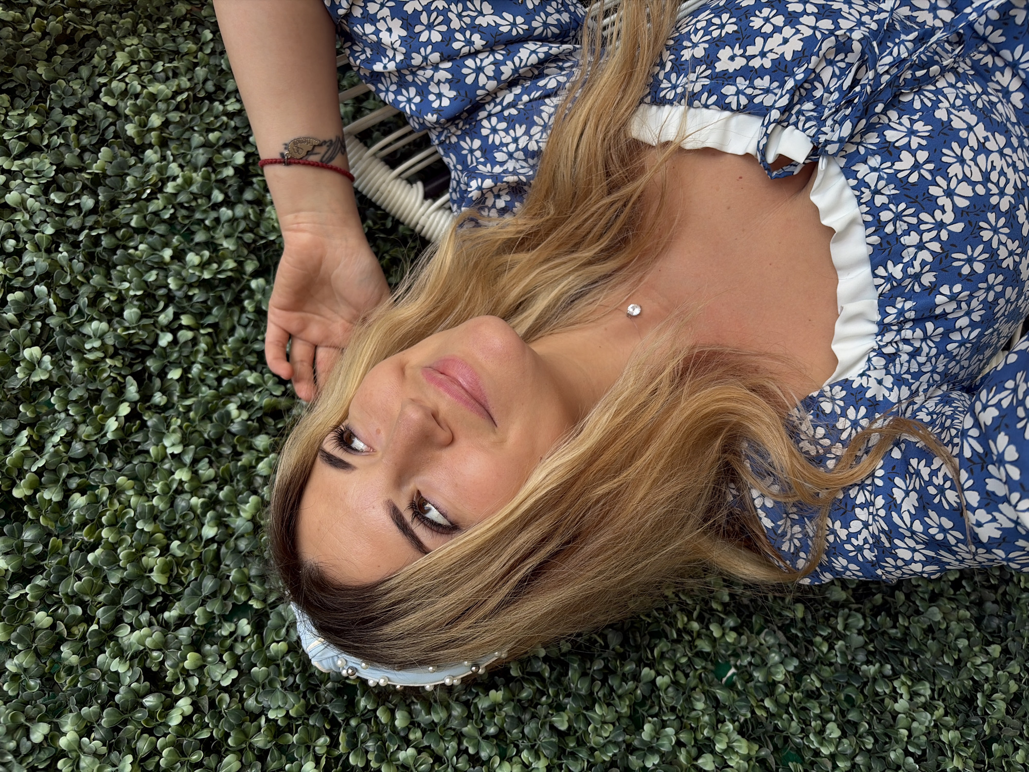 A young woman with blonde hair and a blue floral dress lies on her side against a green foliage wall. She is wearing a pearl-embellished headband and a delicate silver necklace with a single clear stone. Her right arm is raised, with her hand resting on the back of her head. A red string bracelet is visible on her left wrist. She is looking up and to her right with a slight smile. The background is a dense wall of artificial greenery, characteristic of many modern outdoor seating areas in tourist destinations like Budva, Montenegro.
