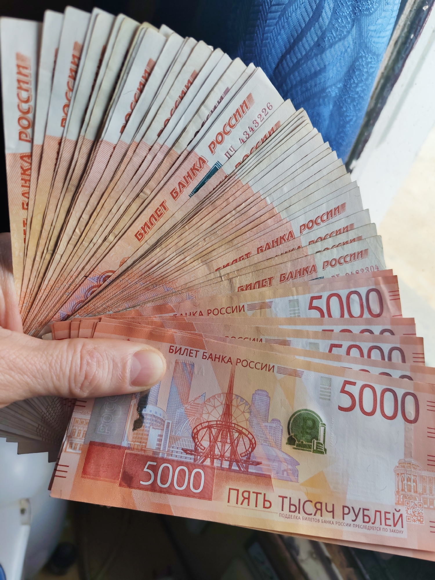 A hand is holding a fan of Russian 5000-ruble banknotes. The banknotes are orange and white, and feature images of landmarks in Yekaterinburg.