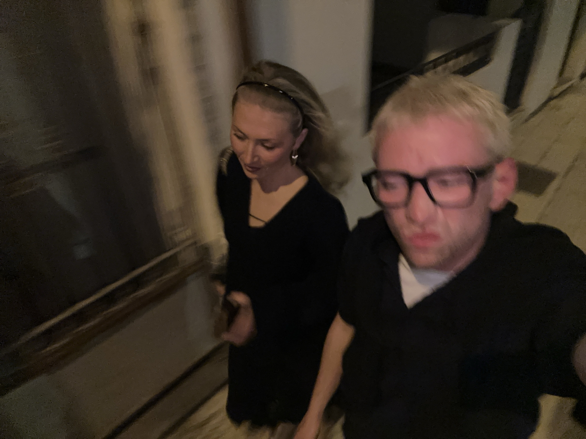 This low-light image captures two individuals in what appears to be an indoor setting, possibly a hallway or staircase, given the blurred vertical lines in the background. The photo exhibits significant motion blur, suggesting either movement by the subjects or the camera, possibly taken as a selfie or video by the person on the right.
The subject on the left is a woman with blonde hair, wearing a dark headband, earrings, and a dark V-neck top or dress. She is looking downwards with a soft expression, and a dark, rectangular object, possibly a phone, is visible in her left hand, which is also blurred.
The subject on the right is a man with light blonde hair and black-framed glasses. He is wearing a dark, collared shirt over a white undershirt. His face is closer to the camera and appears somewhat distorted and blurred, suggesting he might be holding the camera. His expression is difficult to discern clearly due to the blur but appears slightly strained or surprised.
The overall lighting is dim, indicating either nighttime or a poorly lit interior space. The background is indistinct and blurry, making it difficult to identify specific architectural details, but some vertical elements suggest walls or railings. No specific visual cues confirm the provided location of Lipari, Italy, within the image itself. There is no visible text.