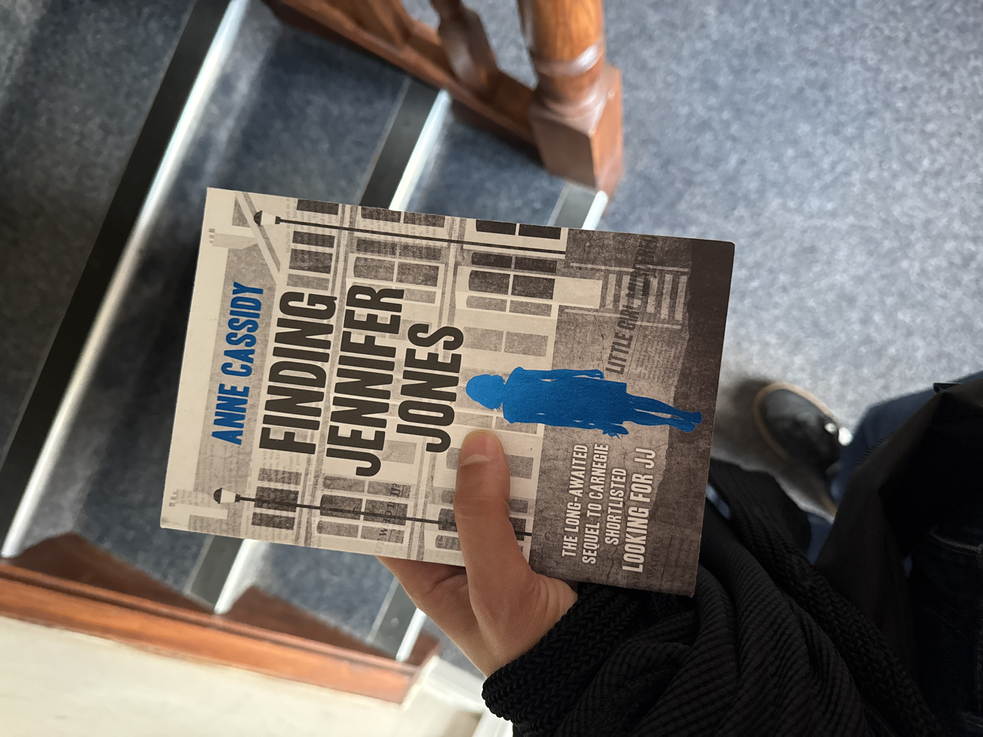 A person is holding an open book titled "FINDING JENNIFER JONES" by ANNE CASSIDY. The book cover features a silhouette of a woman in blue and a cityscape graphic. The person is wearing a black knitted garment over a dark blue top and blue jeans. They are standing on carpeted stairs, with wooden handrails visible on either side. The background shows a carpeted floor and wooden banisters. There is no visible indication of the city of Great Yarmouth, United Kingdom.