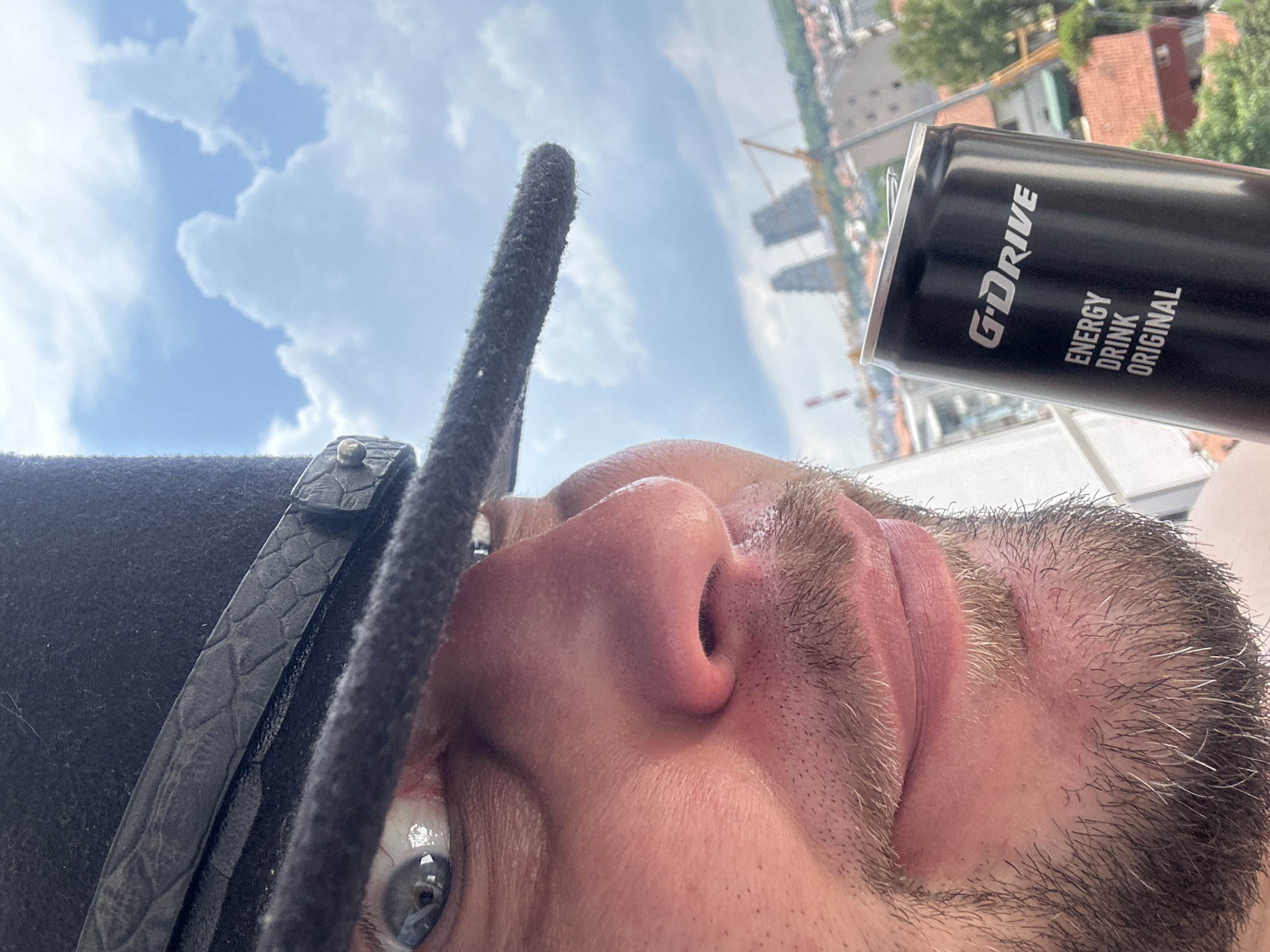 The image is a close-up, fisheye-lens perspective of a person's face and a can of G-DRIVE ENERGY DRINK ORIGINAL. The person is wearing a black hat with a textured band. Their eye is visible, and they appear to be looking upwards. The background shows a cityscape with buildings and cranes, suggesting an urban setting. The sky is visible with clouds, indicating it is daytime. The scene is outdoors, possibly on a balcony or elevated platform given the city view. The angle and lighting suggest a selfie taken in natural light. There is no strong indication of a specific emotion. The can of energy drink is black with white text.