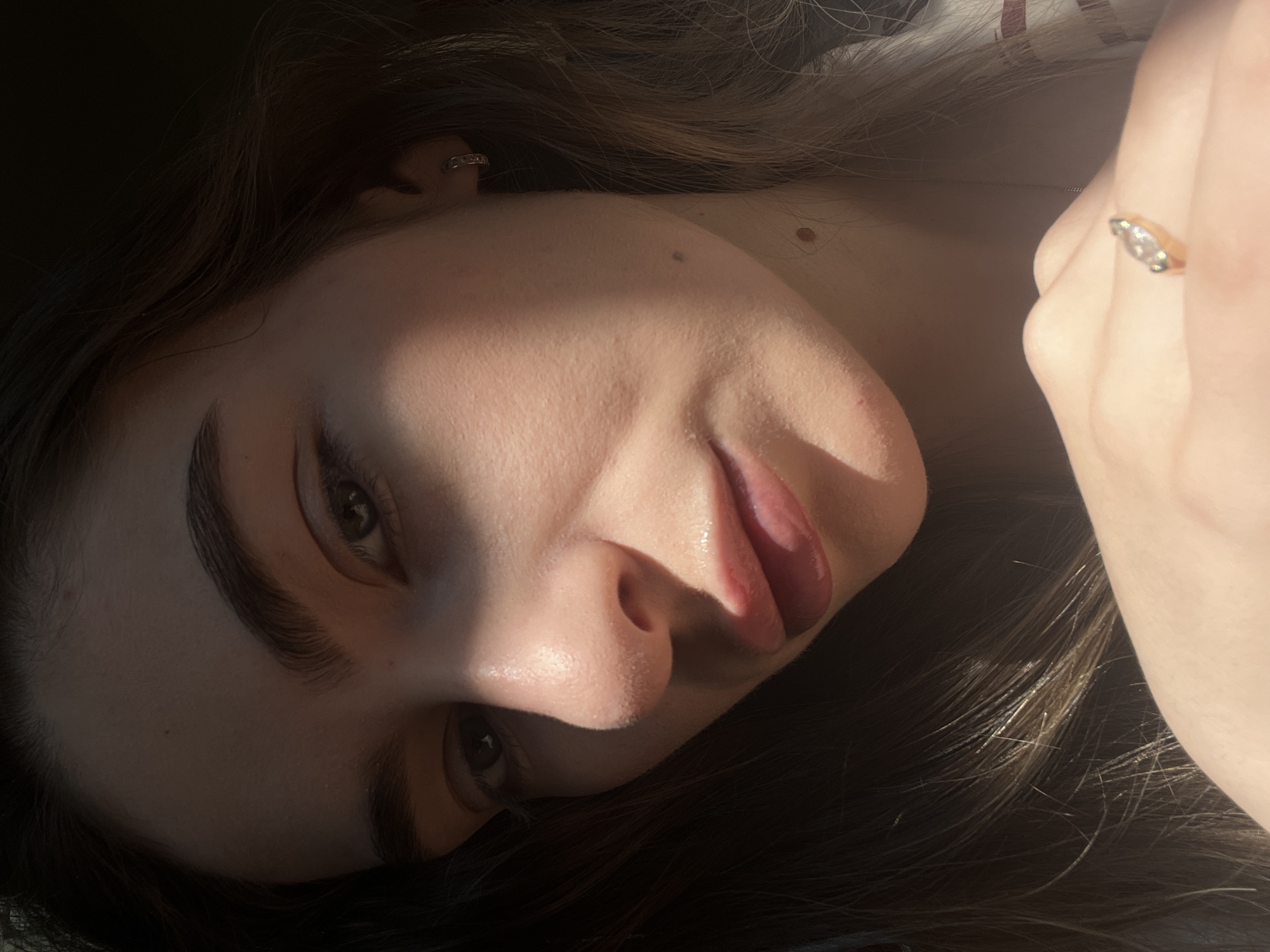 A young woman with brown hair and fair skin is lying down, her face tilted towards the camera. She has thick eyebrows, and her tongue is slightly protruding from her mouth. Light and shadow create a contrast across her face, highlighting her features. A ring is visible on a finger near her face. The location and city are unknown.