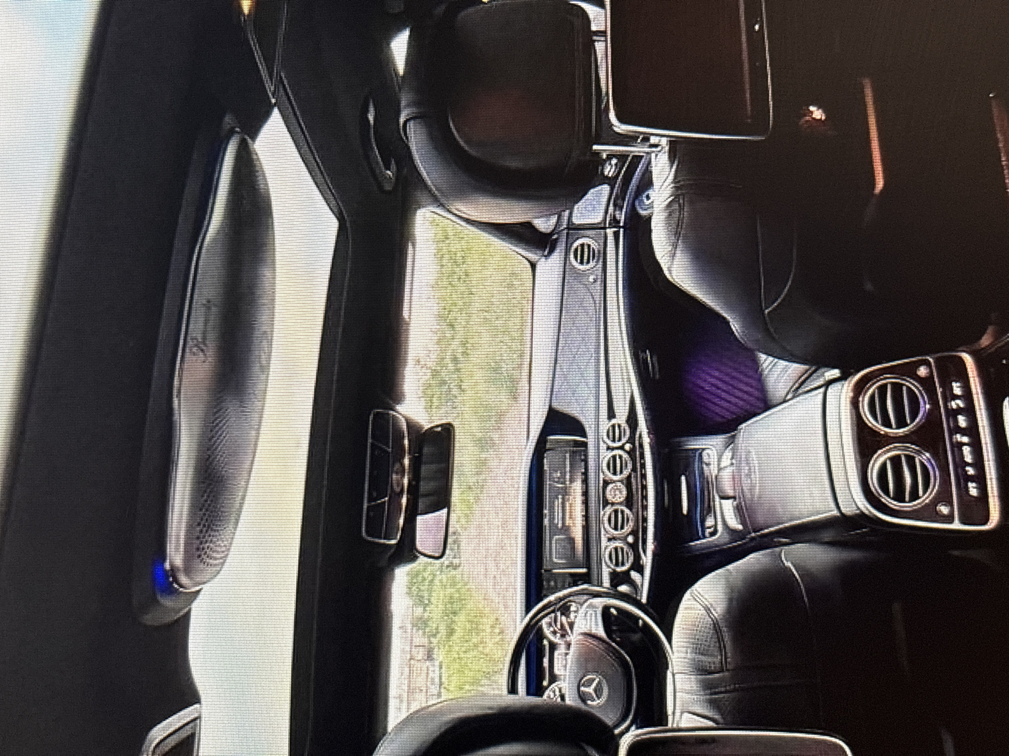 The image shows the interior of a luxury car, likely a Mercedes-Benz S-Class, viewed from the rear passenger seats. The car has a panoramic sunroof, black leather upholstery, and a modern dashboard with a large infotainment screen. The front seats and steering wheel are visible, as is the center console. The exterior view through the windows suggests a rural or suburban setting with a dirt road and vegetation. There are no people visible inside the car. The image appears to have been taken with a low-resolution camera, resulting in a pixelated appearance. The scene is static, with no discernible action or interaction taking place. There is no information within the image to suggest the location is Moscow, Russia.
