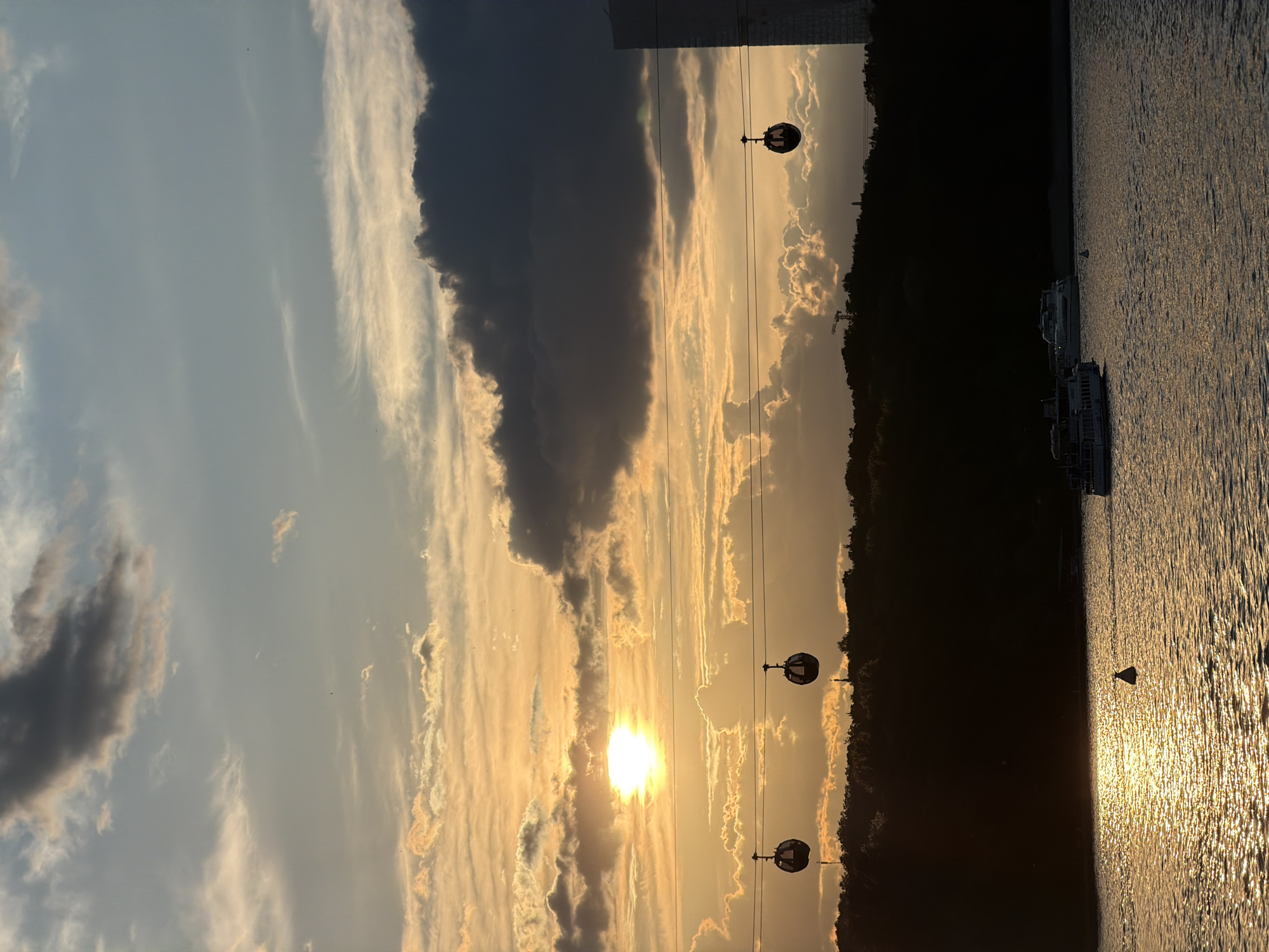 This landscape image captures a vibrant sunset over a wide body of water, likely the Moskva River in Moscow, Russia, given the context. The sun, a bright golden orb, is low on the horizon, casting a brilliant reflection across the rippling water surface in the foreground.
The sky occupies the upper portion of the frame, painted with warm, golden hues near the horizon where the sun sets, transitioning to lighter blues and grays higher up. Various clouds are scattered across the sky: some are dark and voluminous, while others are elongated and wispy, catching the golden light.
Stretching across the midground are several cable car gondolas, suspended from wires. Three spherical, dark-colored gondolas are clearly visible, contrasting with the bright sky behind them. They appear to be part of an active transportation system, like the Moscow Cable Car.
Below the cable cars and across the horizon line, a dark silhouette of land, densely covered with trees, creates a stark contrast against the radiant sky. A small construction crane is faintly visible among the trees in the distance. On the water, several white boats or small ships are moored or slowly moving, adding to the sense of an active waterway. A dark, triangular buoy or marker floats in the middle of the glowing water.
The overall scene conveys a serene and picturesque evening, with the dramatic sunset light being the dominant feature, highlighting both natural elements and man-made structures. There is no visible text in the image.