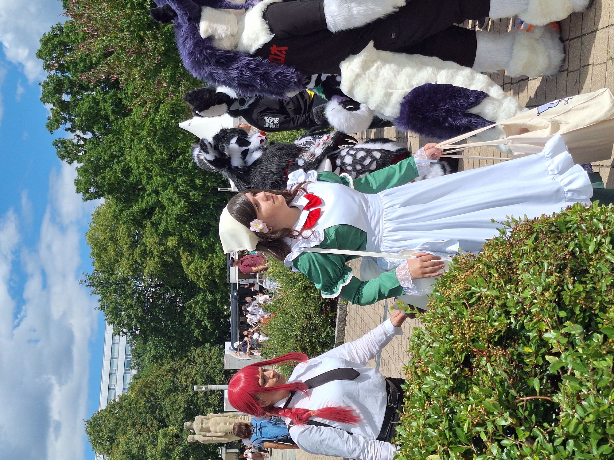 In Chemnitz, Germany, at the Staatstheater square, a group of individuals are seen walking outdoors on a sunny day. The foreground features a person in a maid costume, wearing a green and white dress with a white head covering and a flower. To their left, a person with bright red hair is dressed in a white shirt and black tie. Behind them, several people are dressed in fursuits, with one in a black and white wolf-like costume, another in a black costume with a white furry chest and paws, and a third in a dark purple and white furry costume. The background shows trees, a building, and a statue. The ground is paved with light-colored stones.
