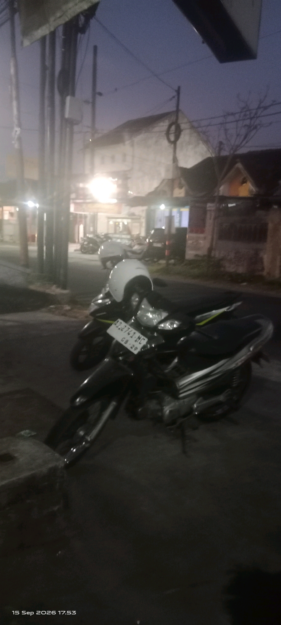 The image shows two motorcycles parked on a street in Merbung, Indonesia at dusk. The motorcycle in the foreground is black with a white helmet resting on its seat. A license plate with the characters "AD.2741.MC 08.29" is visible. The second motorcycle, also dark-colored, is parked behind the first. The background features buildings, streetlights, and utility poles, suggesting an urban or semi-urban setting. The sky is a deep blue, characteristic of twilight. The overall scene is dimly lit.