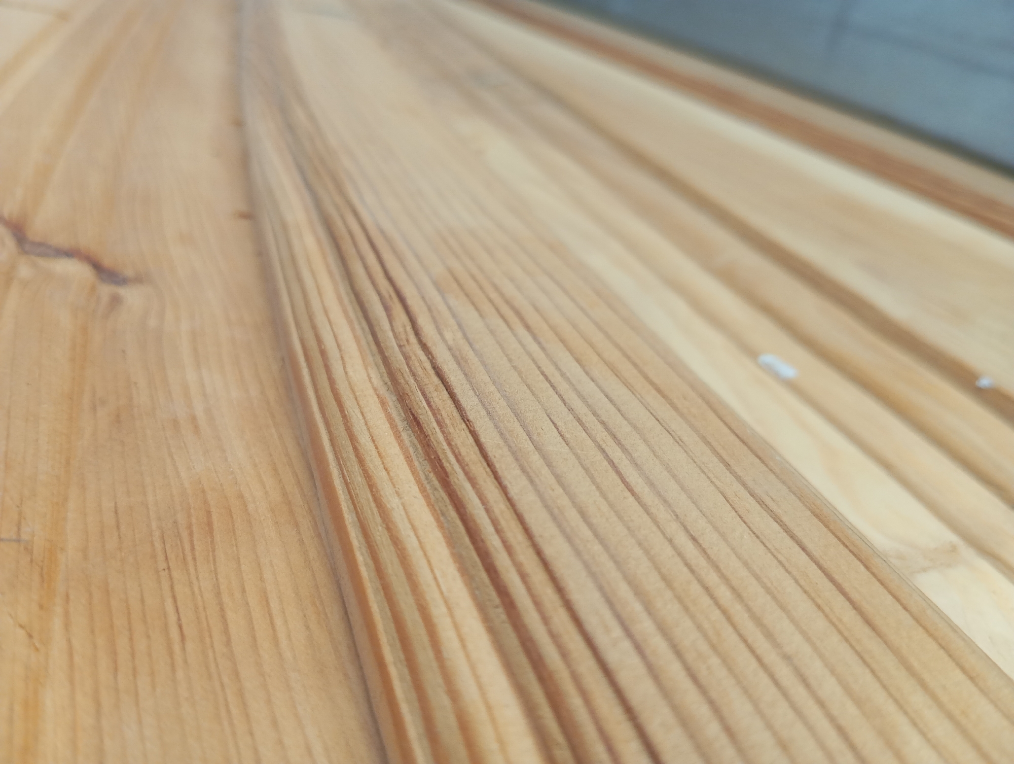 The image is a close-up, high-angle shot of a wooden surface. The wood has a light brown color with prominent grain patterns that run diagonally from the bottom left to the top right. The surface appears to be made of several planks, with visible seams between them. The wood has a smooth, finished texture. The background is out of focus, showing a blurred gray wall on the right side. The lighting is natural and appears to be coming from the top left, creating subtle shadows that highlight the wood grain. There are no people or animals visible in the image.
