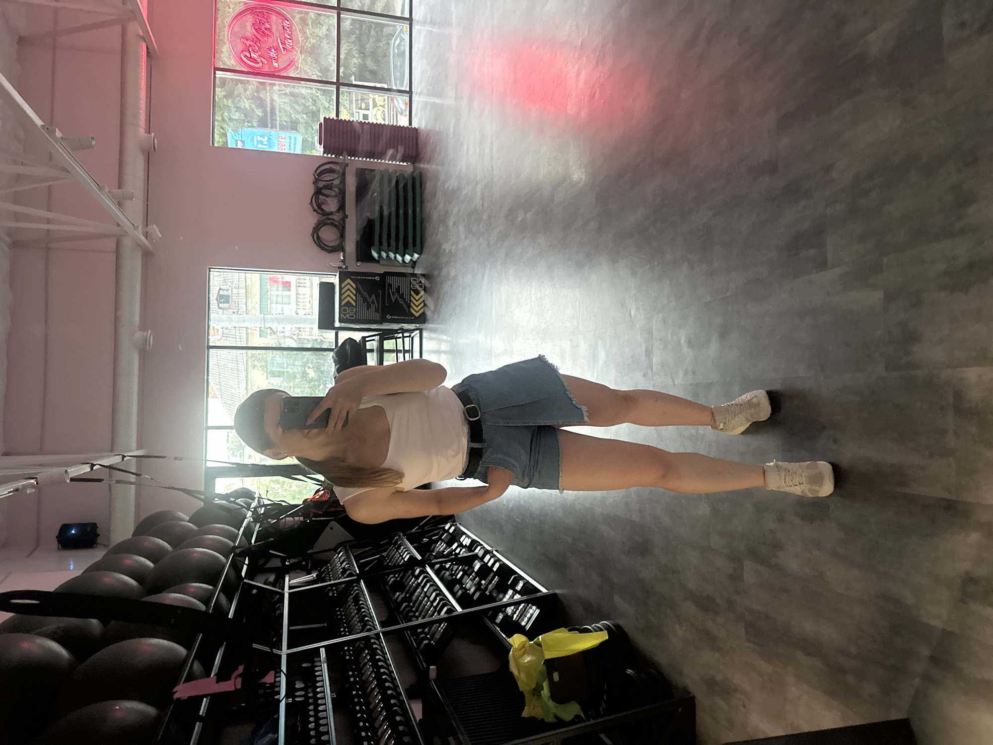 A young Caucasian woman with blonde hair pulled into a ponytail is taking a mirror selfie in what appears to be a gym. She is wearing a white tank top, denim shorts with an asymmetrical hem, a black belt, and light-colored sneakers. She is standing in front of a wall of dumbbells and exercise balls. To her right, there are large windows offering a view of buildings and trees, suggesting an urban setting. The floor is a dark, wood-effect laminate. The lighting in the gym is bright, with a pink neon sign visible in the background. The presence of gym equipment and the overall aesthetic suggest this is a fitness facility. The image provides no specific clues or markers to definitively place it in the city of Ternopil, Ukraine.