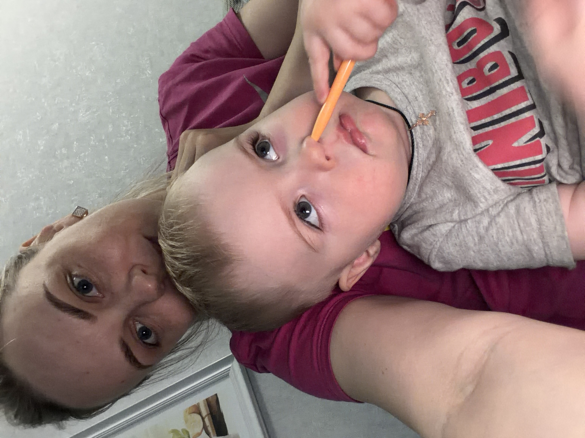 An eye-level, rotated candid shot features a woman and a young child in an indoor setting, likely a home in Votkinsk, Russia. The image is rotated 90 degrees clockwise.
The woman, on the left, has light-colored hair, pulled back, and blue eyes. She wears a dark pink or magenta top and a sparkling ring on her right hand. Her expression is neutral and direct towards the camera.
The child, held by the woman, is positioned above her. The child has light blonde hair and blue eyes, looking towards the camera or slightly beyond it. They are wearing a grey t-shirt with large red letters outlined in black, which appear to spell "NIKE" or a similar brand, though partially obscured and rotated. A small, gold-colored cross hangs from a black string around the child's neck. The child is holding an orange, slender object, possibly a spoon or a small tool, and has it positioned near their nose and mouth. The child's expression is one of focused curiosity.
The background is a light-colored, textured wall. A glimpse of a white-framed picture or mirror can be seen in the lower left corner of the frame. The overall atmosphere appears casual and intimate, capturing a moment of interaction between the two subjects.