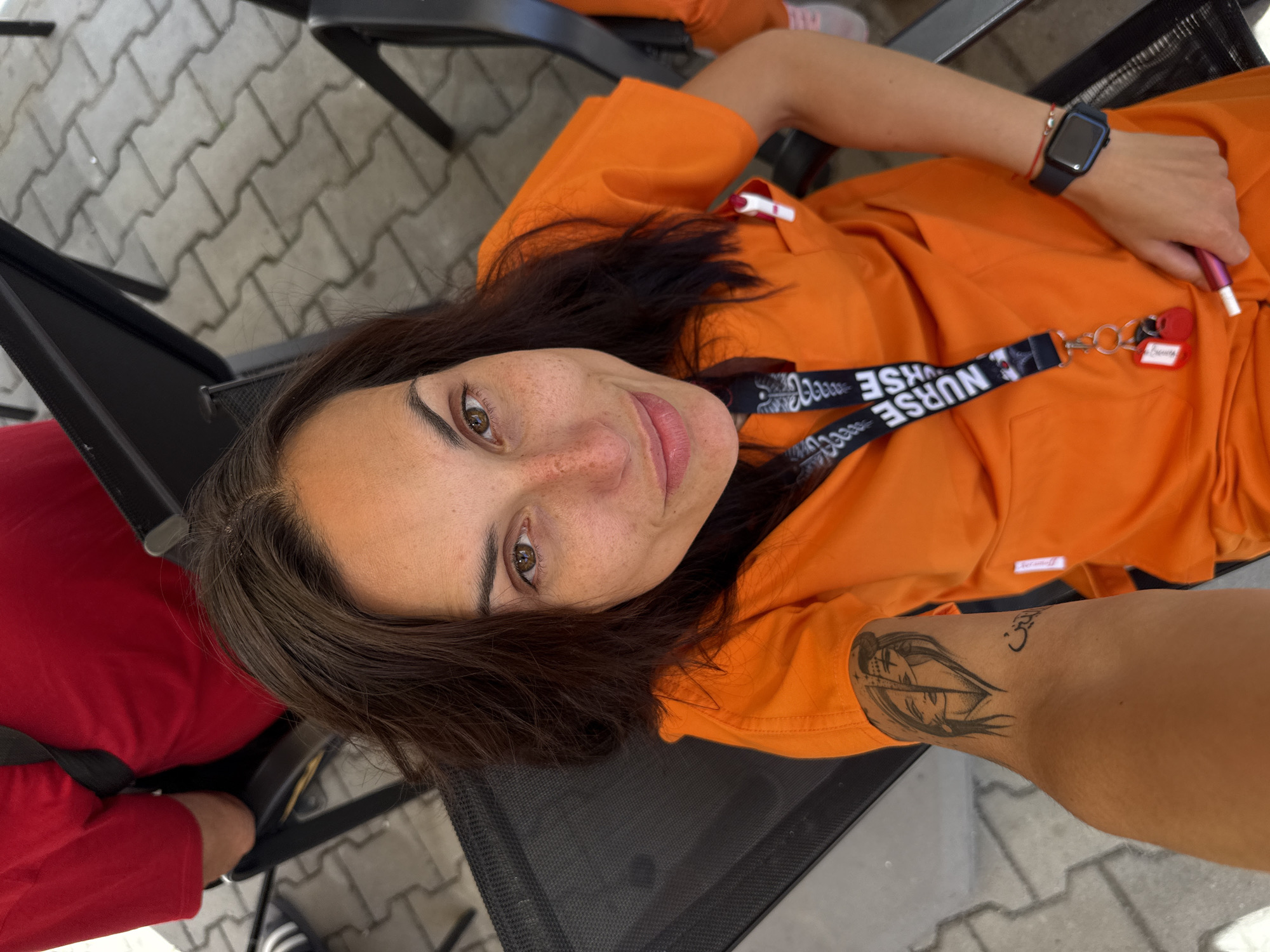 A medium shot captures a woman in bright orange scrubs, sitting in an outdoor setting. She is looking directly at the camera, with a faint smile. Her dark brown hair is loose, framing a face dotted with freckles. A lanyard with "NURSE" printed on it hangs around her neck, with a small red keychain attached. On her left arm, a tattoo depicts two faces, one with a devilish horn, and some Arabic script. She wears a black smartwatch on her right wrist. The background shows paving stones and black chairs, suggesting an outdoor seating area. The scene is consistent with the exterior of a medical facility or a public space in the city of Pleven, Bulgaria.