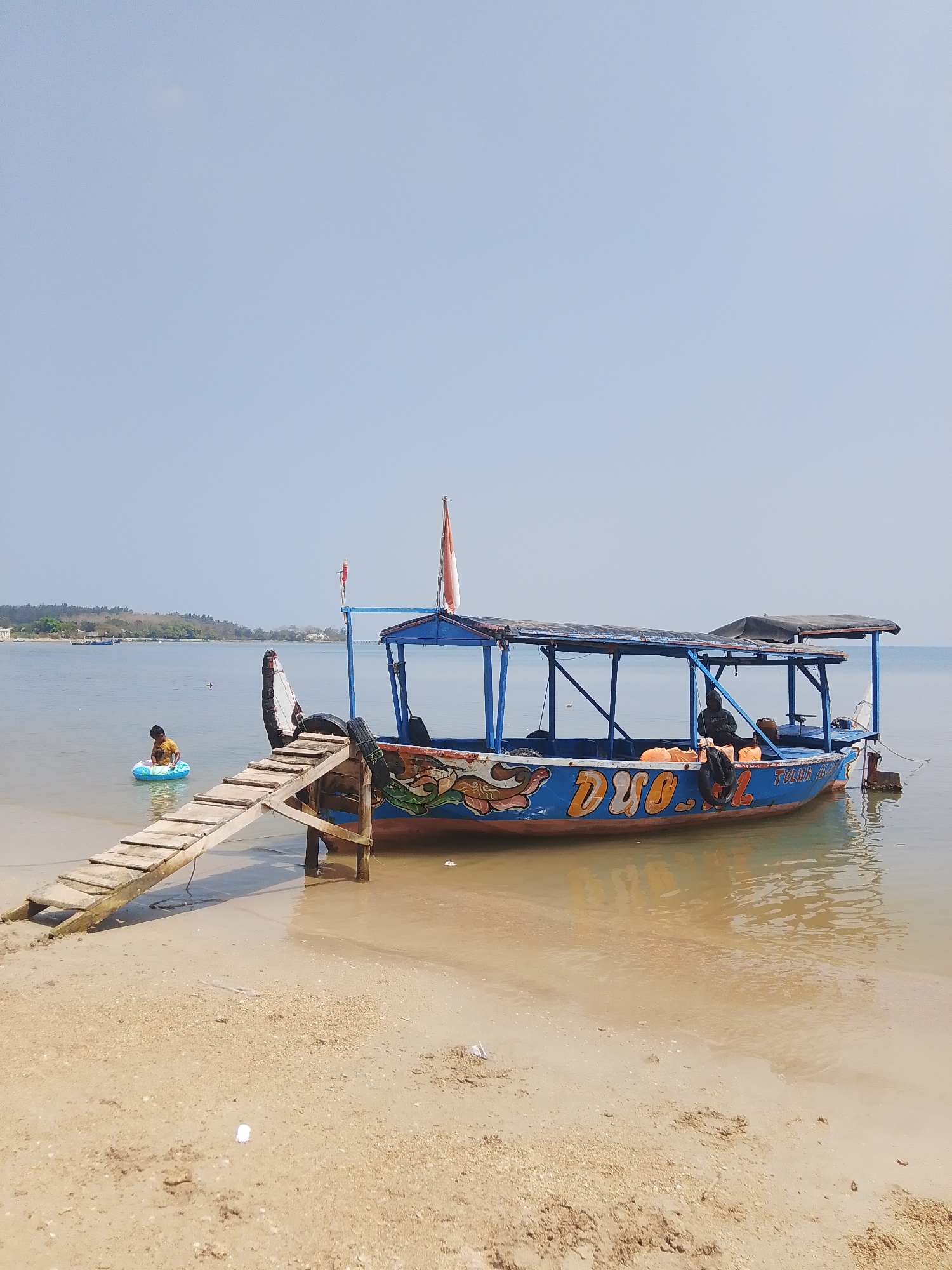 A long, narrow boat with a blue hull and a covered seating area is moored in shallow, calm, tan-colored water near a sandy beach. The boat has colorful decorations on its hull, including stylized orange letters that appear to spell "DUO." Two people are visible on the boat, one sitting towards the back and another further forward. An orange life jacket is visible on the back of the boat. A wooden ramp extends from the boat to the water. To the left of the boat, a young child wearing a yellow shirt is sitting in a blue inner tube in the water, near the shore. The sky is clear and pale blue. In the background, a distant, tree-covered coastline is visible. The scene appears to be a tranquil day at the beach in Pejagan, Indonesia.