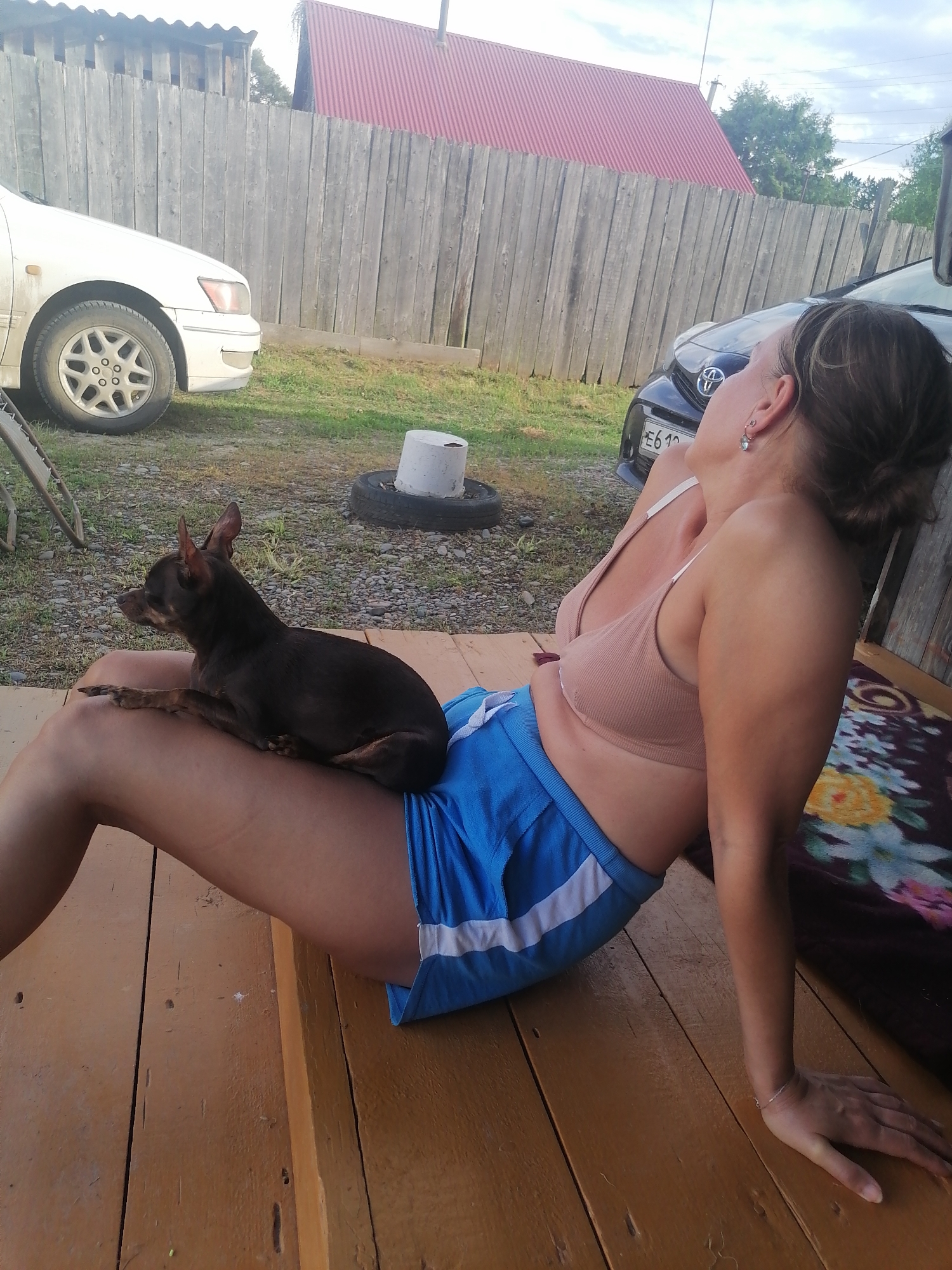 A woman with brown hair, wearing a beige bralette and blue shorts, is sitting on a wooden porch with a small, dark brown dog on her lap. She is looking upwards and to the right. A wooden fence, a car, and a red-roofed building are visible in the background. The scene appears to be in a rural or suburban area, consistent with the location of Novopokrovka, Russia.