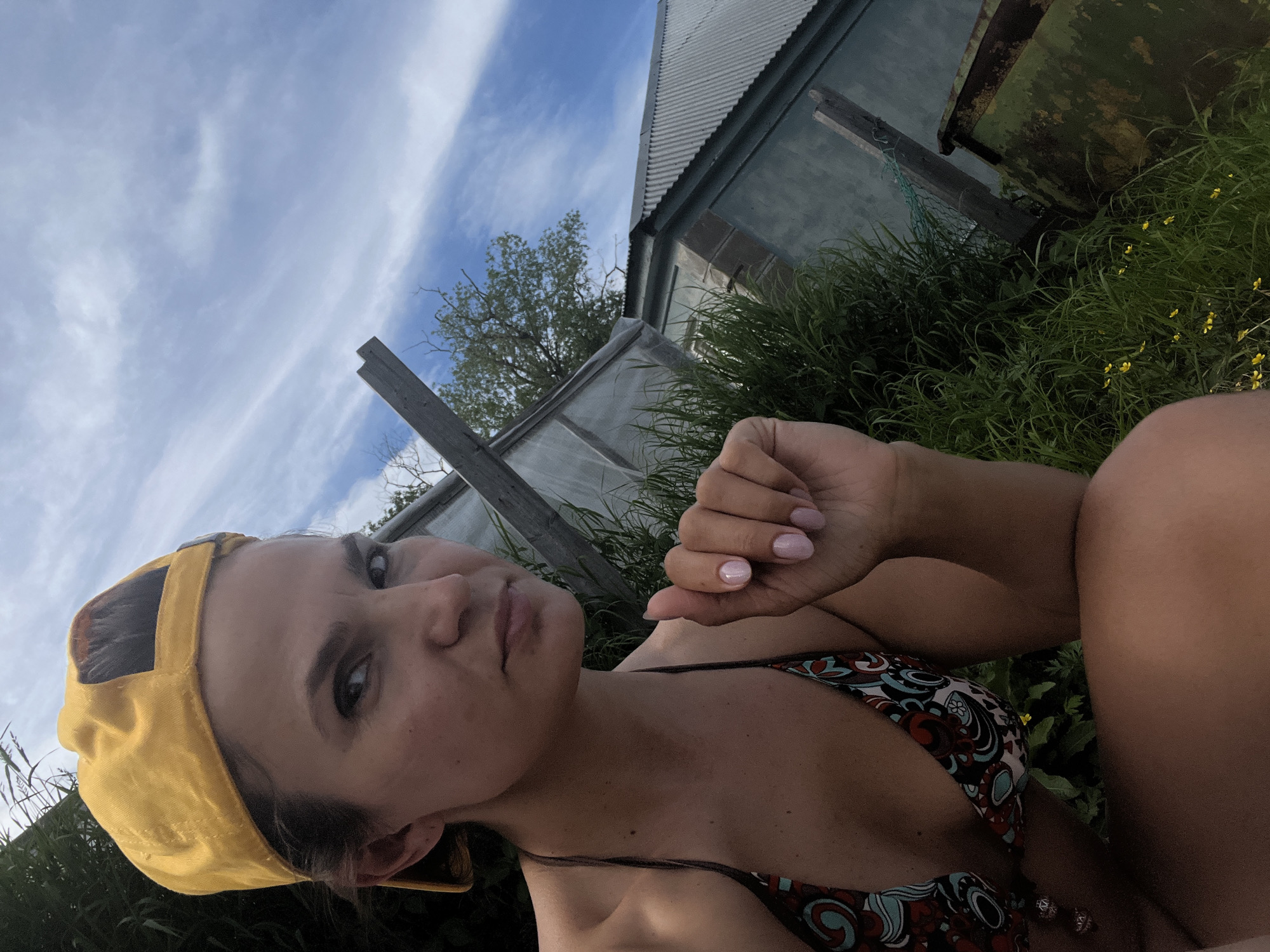 A woman wearing a yellow baseball cap and a patterned bikini top is lying down outdoors, with her face turned towards the camera. She has dark hair and is looking directly at the viewer with a neutral expression. Her right hand is raised, showing manicured nails. She is surrounded by tall green grass and some small yellow flowers. In the background, there is a building with a corrugated metal roof, a wooden structure, and what appears to be a greenhouse or similar transparent-covered structure. The sky above is blue with white, wispy clouds, suggesting it is daytime and the weather is fair. The scene is likely a rural or semi-rural setting, possibly a backyard or a field. There is no visible text in the image.