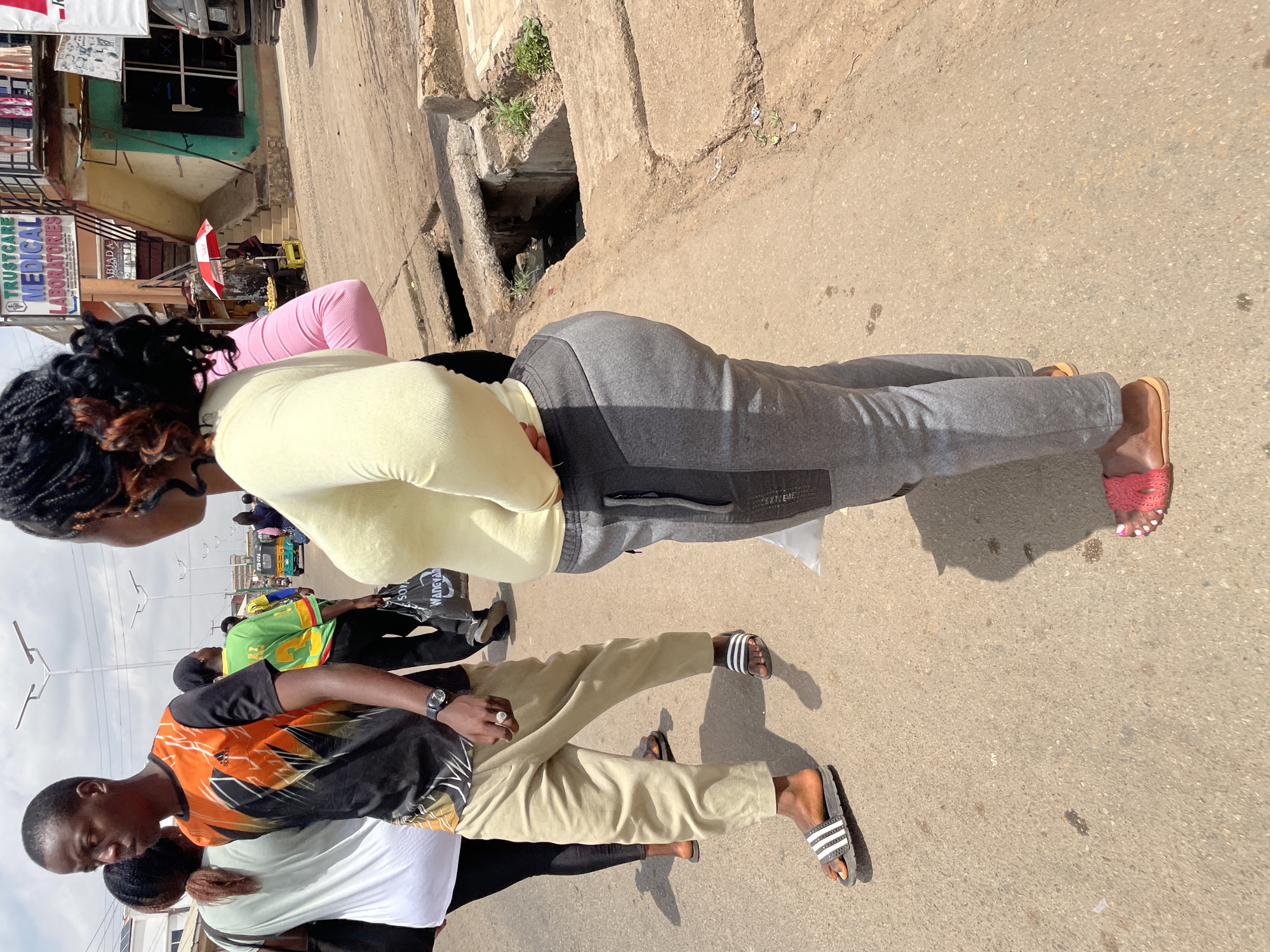 The image shows a street scene in Jos, Nigeria. The ground is paved and cracked. A storm drain is visible along the roadside. Several people are walking on the street. One person is wearing a yellow shirt, pink pants, and gray sweatpants, and sandals. Another person is wearing an orange and black shirt and tan pants, with Adidas sandals. A sign for Trustcare Medical Laboratories is partially visible in the background.