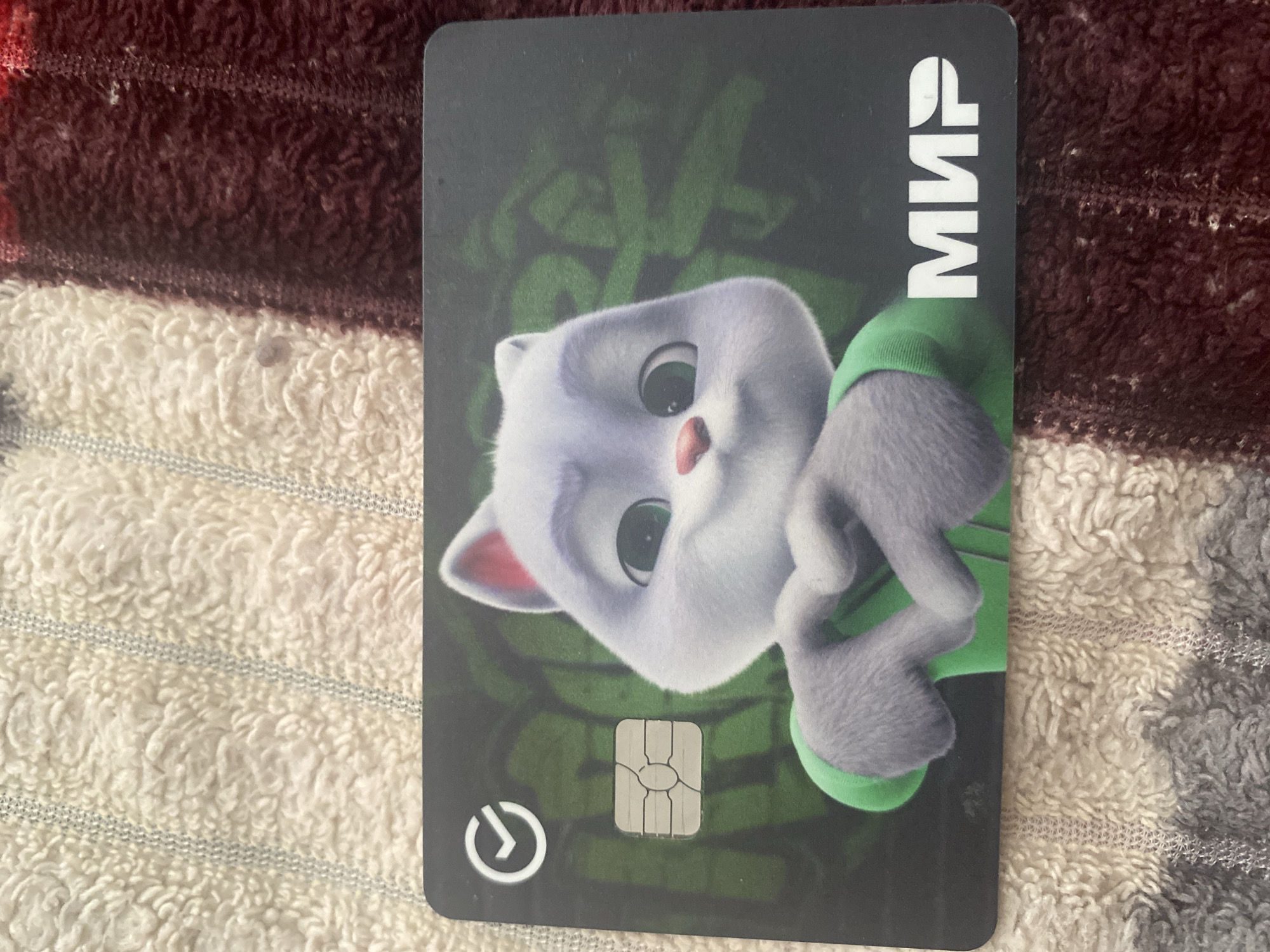 A close-up shot shows a debit card with a cartoon cat character making a heart shape with its paws. The card features the "MIR" payment system logo and a circular checkmark icon. The card is placed on a textured surface, possibly a blanket or towel, with alternating beige and dark red striped patterns. There is no discernible location in Kizilyurt, Russia, depicted in the image.