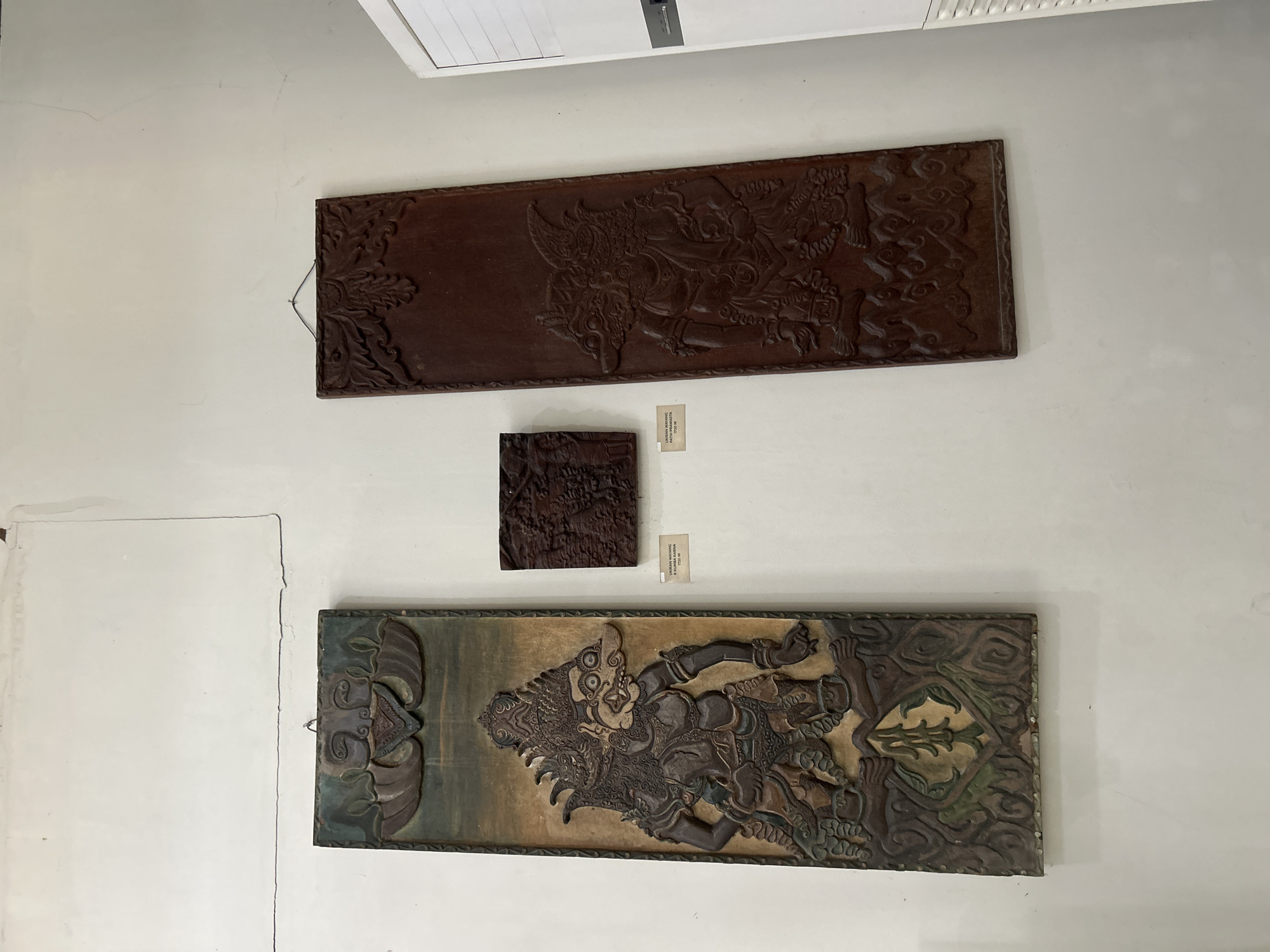 This image depicts three carved wooden panels displayed on a white wall, likely within a museum or cultural heritage site in Cirebon, Indonesia. The largest panel on the left is a vertical rectangular carving of a grotesque figure with an elaborate headdress, detailed armor, and clawed hands, rendered in muted earth tones and green. Below the figure is an ornamental shield-like motif. To its right is a smaller, square-shaped carving with intricate, dense relief patterns, appearing to be a darker wood. Further to the right is another tall, vertical rectangular panel, also featuring a carved figure in a similar style to the left panel but with a darker, reddish-brown hue and less distinct coloration. All three panels are hung with simple wire. A partially visible white air conditioning unit is present on the far right of the frame. Small informational plaques are visible beneath the two smaller panels. The wall exhibits some minor discoloration and subtle cracks, suggesting an indoor environment with aged plaster.