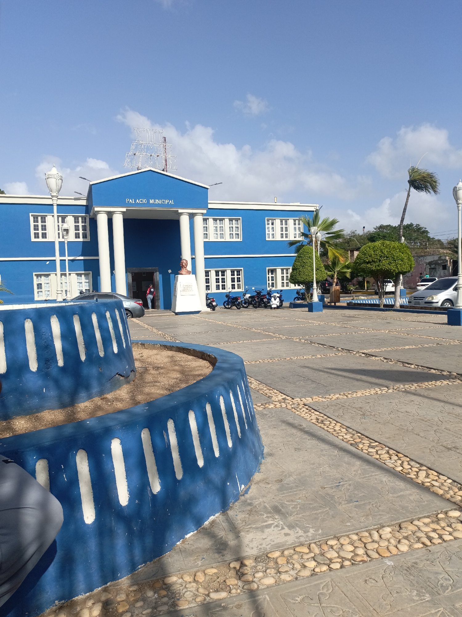 The image shows the Palacio Municipal in Juan Griego, Venezuela. The building is a vibrant blue with white trim and columns, topped with the words "Palacio Municipal." A small, two-tiered blue and white fountain sits in the foreground. The plaza in front of the building is paved with a combination of concrete and decorative stones. A bust statue stands near the entrance. Several parked motorcycles and cars are visible. The sky is blue with scattered clouds. Palm trees and other greenery surround the plaza. A person is entering the building.