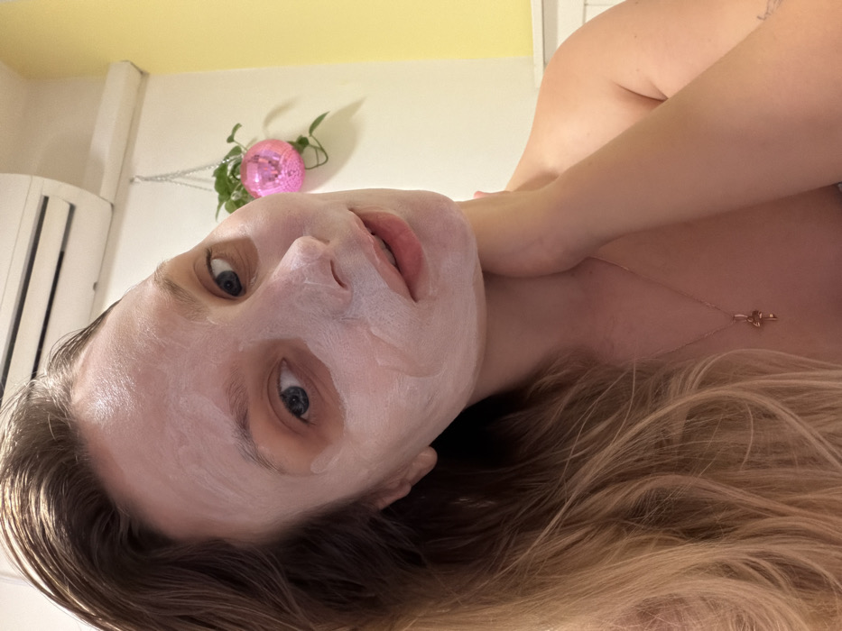 The image is an upward-angled, close-up portrait of a young adult female, likely lying down, engaged in a self-care activity. Her face is covered in a thick, white face mask, with her blue eyes looking directly at the camera and her lips slightly parted. She has long, blonde hair that spreads out around her head and shoulders, and a delicate, gold-colored necklace with a small pendant is visible on her collarbone.
The background shows a light-colored, possibly yellowish-beige wall. To the far left, a white air conditioner unit or similar appliance is partially visible. Directly behind her head, a small, pink disco ball hangs on the wall, adorned with green leaves, possibly from a plant or artificial foliage. The lighting suggests an indoor setting. The composition indicates a self-portrait, capturing a personal moment. This scene is set indoors, within a residential context in Tel-Aviv, Israel. There is no visible text in the image.