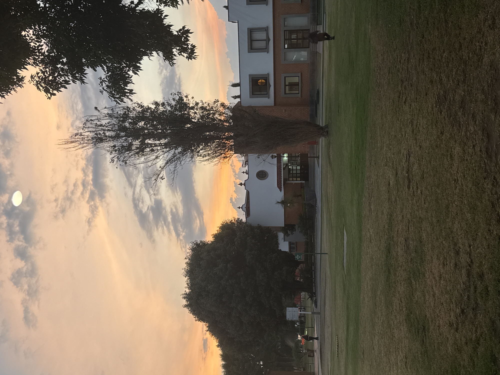 The image depicts a building at sunset in San Andrés Cholula, Mexico. The sky is filled with clouds and a gradient of orange, yellow, and grey colors. A bright, round object, possibly the moon, is visible in the upper left. Trees are silhouetted against the sky in the foreground and middle ground. A two-story building, painted white with red brick details, is situated in the center of the image. Its windows are visible, and a figure is standing on the grass near the building. A large grassy field occupies a significant portion of the scene, with its texture noticeable. The scene appears to be a park or a public space.