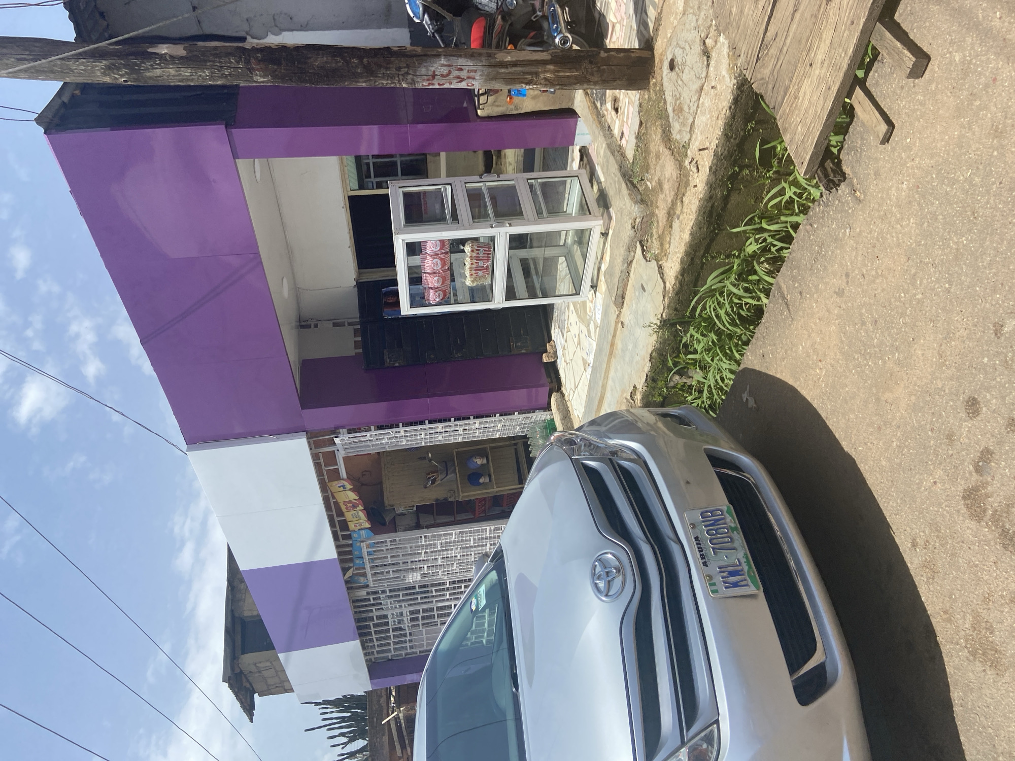 A silver Toyota Sienna minivan, with license plate KWL-708NB, is parked in front of a store in Jos, Nigeria. The store has a purple and white facade. In front of the store is a glass display case with popcorn containers inside. To the right of the store, a wooden pole stands next to a motorcycle. The ground is paved with concrete and some areas are covered with dirt and grass. The sky is cloudy.