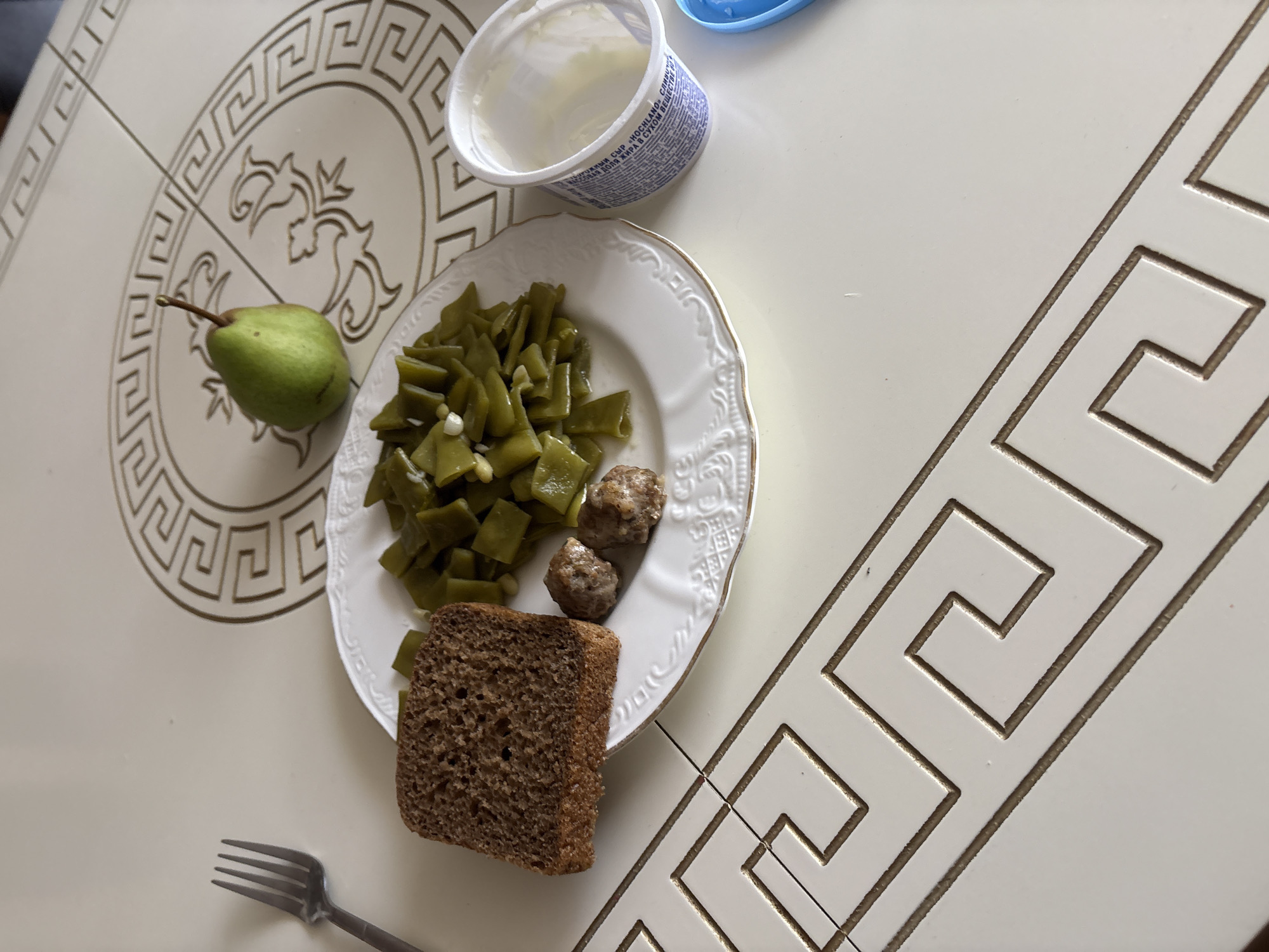 The image displays a meal laid out on a white, patterned table. On a white plate with a gold rim, there are chopped green beans, two small meatballs, and a slice of dark bread. To the right of the plate, a small container of what appears to be yogurt is visible. A single green pear rests on the table to the upper right of the plate. A silver fork is positioned to the left of the plate. The table itself has a decorative border pattern. The scene is lit from above, casting soft shadows. This image does not contain enough information to determine its location within the city of Georgiyevsk, Russia.