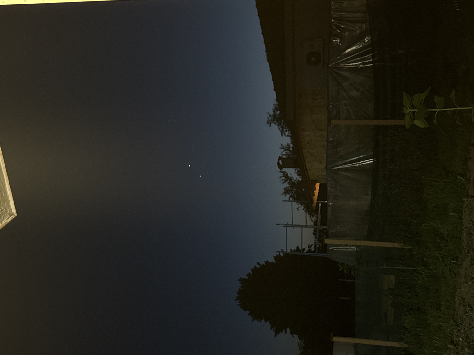 The image captures a nighttime scene in Rogosh, Bulgaria, likely from an elevated vantage point such as a balcony, as suggested by the angled, illuminated ceiling edge in the top portion of the frame. The sky is a deep, dark blue, characteristic of twilight or nighttime. Three bright celestial bodies, possibly planets or stars, are visible in the upper half of the sky. The lower half of the image depicts a residential area at dusk or night. A dark, silhouetted tree stands to the left. In the middle ground, scaffolding is erected in front of a building with a tiled roof and a chimney. A portion of the building's exterior wall is visible, along with an air conditioning unit. A transparent plastic sheeting barrier runs horizontally across the mid-ground. In the foreground, tall grass and a young plant, possibly a sunflower, are visible. The overall lighting suggests artificial illumination from the building and potentially streetlights, casting a warm glow on the structures.
