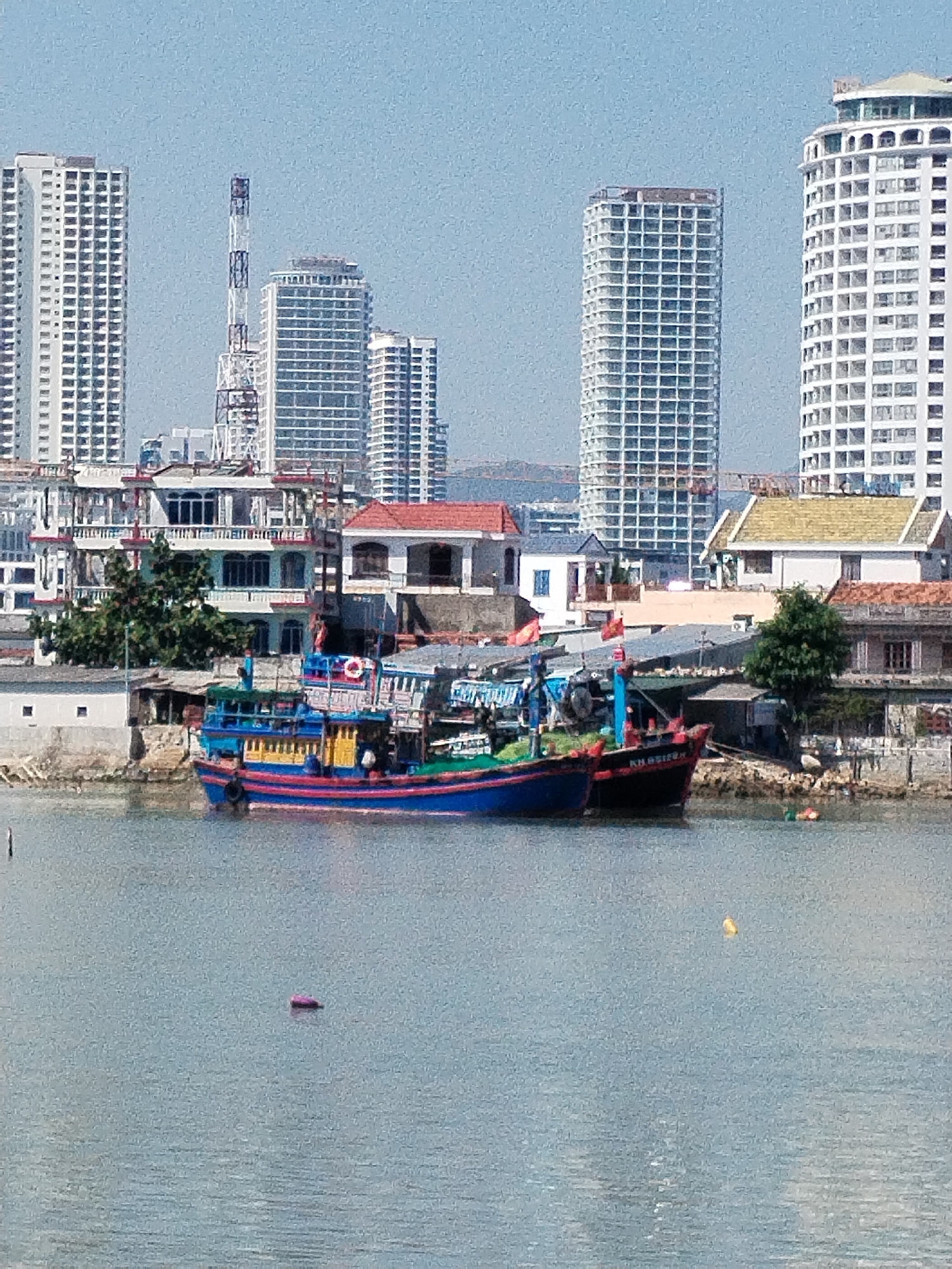 The image shows a waterfront scene in Bac Nha Trang Ward, Vietnam, with two fishing boats docked near the shore. The boats are painted in bright colors, including blue, red, green, and yellow. One boat has a life preserver visible on its side. Several people can be seen on or near the boats. Behind the boats, the shoreline is lined with buildings of varying sizes and architectural styles, including residential houses and larger structures, some with red tile roofs. In the background, several high-rise buildings dominate the skyline, creating a modern urban backdrop. There is also a tall communications tower visible. The water in the foreground appears calm, with a few small objects floating on the surface. The sky is clear and blue, indicating a sunny day.