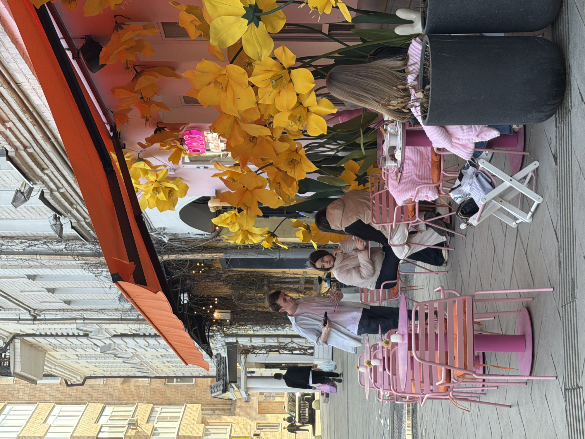 An outdoor cafe is depicted on a paved city sidewalk on Ulitsa Bol'shaya Dmitrovka, Moscow, Russia. The cafe facade, colored light pink, is extensively decorated with large, artificial yellow flowers resembling daffodils. A neon pink sign displaying "OMG" is mounted on the facade beneath an orange retractable awning. The outdoor seating area comprises multiple small, round, pink tables and matching pink metal slat-back chairs, some equipped with orange seat cushions. Small white vases holding decorative elements are present on the tables.
In the midground, a young man, possibly a cafe staff member, stands facing seated customers. He wears a light-colored unbuttoned shirt over a pink t-shirt and dark pants, holding a dark electronic device (likely a payment terminal or phone) and a lighter, small object in his hands. Three individuals are seated at two pink tables. At one table, two women are present: one wearing a light brown padded jacket looks to her right, while the other is partially obscured. At a table in the foreground right, a person with long, light brown hair is seen from the back, wearing a dark top and draped in a pink blanket. A white cup and saucer are on their table. A small white folding stool next to this foreground table holds a white purse, a dark purse, and other miscellaneous items. Several large, dark gray cylindrical planters are positioned along the cafe's edge, one containing dried vegetation.
The background reveals a city street with a multi-story brick building on the left. A pedestrian in a dark jacket carrying two light-colored plastic bags walks away from the camera. A dark-colored SUV or jeep is parked on the street, and two additional figures are visible further down.