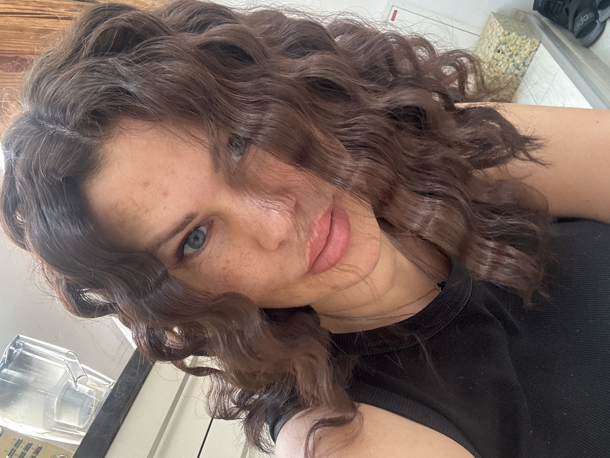 The image is a close-up, selfie-style shot of a woman with medium-length, wavy brown hair and blue eyes. She has visible freckles on her face. She is wearing a black, sleeveless top and a silver necklace. The background appears to be indoors, with a kitchen appliance and a piece of foam material visible. There is no indication of the city of Katowice, Poland in the image.