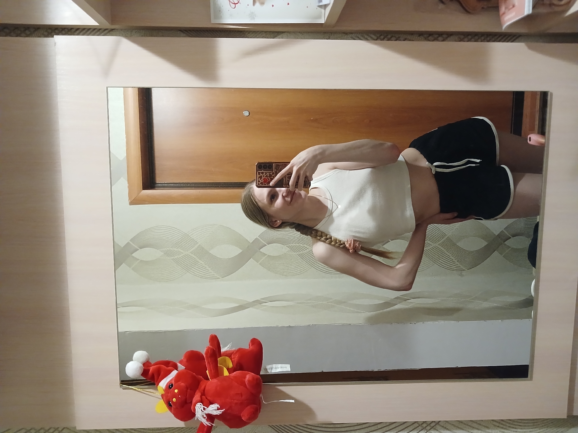 The image depicts a woman taking a selfie in a mirror. She is wearing a white tank top and black shorts with white trim. Her hair is blonde and braided. The mirror reflects a wooden door, a patterned ceiling, and a red stuffed animal. The location is in Chaikovsky, Russia.